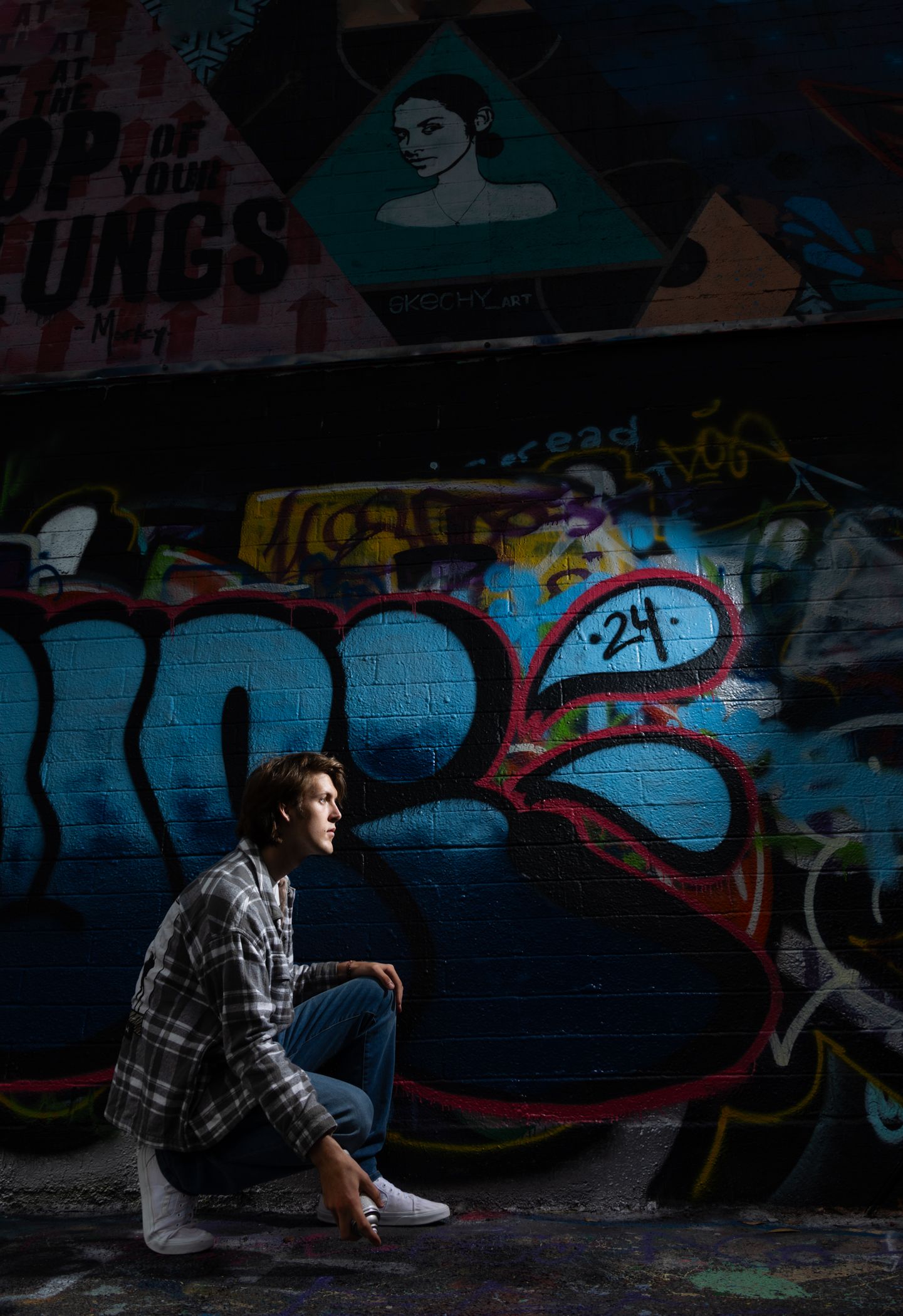 Young man sitting against a vibrant graffiti wall in an urban alley, wearing a plaid shirt and jeans.