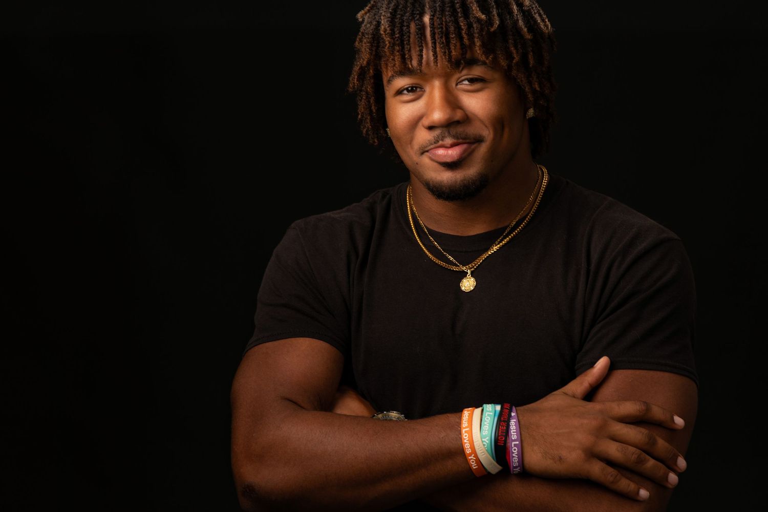 Smiling young Black man in black shirt with colorful bracelets posing against dark background.