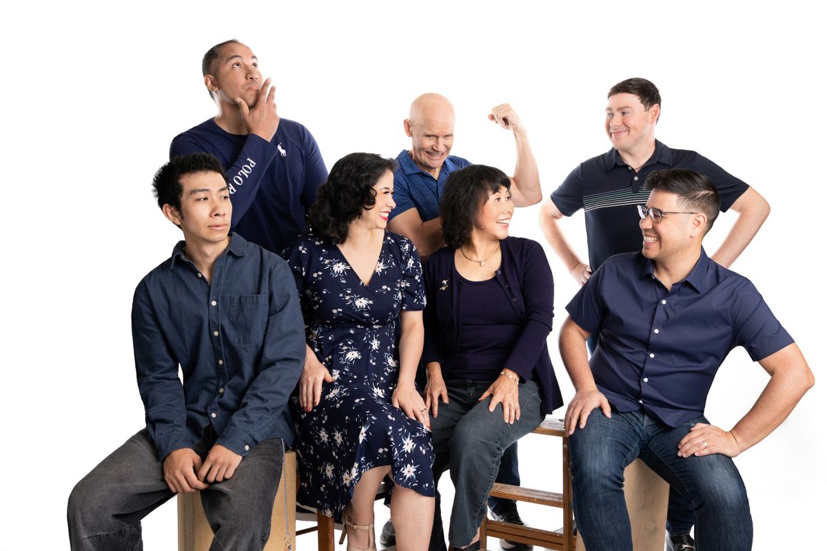 Seven diverse friends pose playfully in navy outfits for a fun group portrait against a white background.