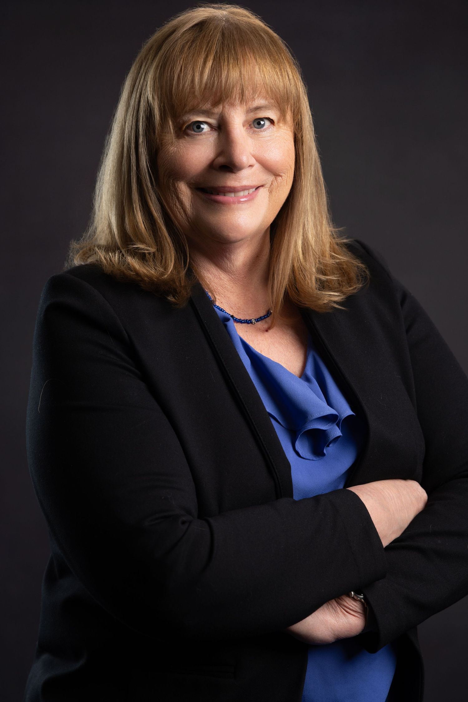 Professional woman in black blazer and blue blouse smiling with arms crossed against dark background.