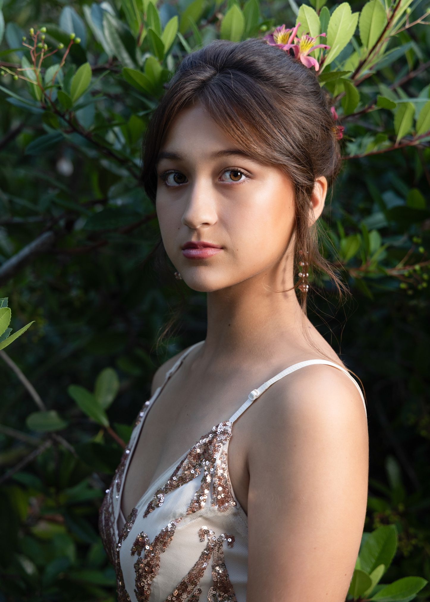 Young woman with floral hair accessories wearing a gold sequin dress posing among green garden foliage.