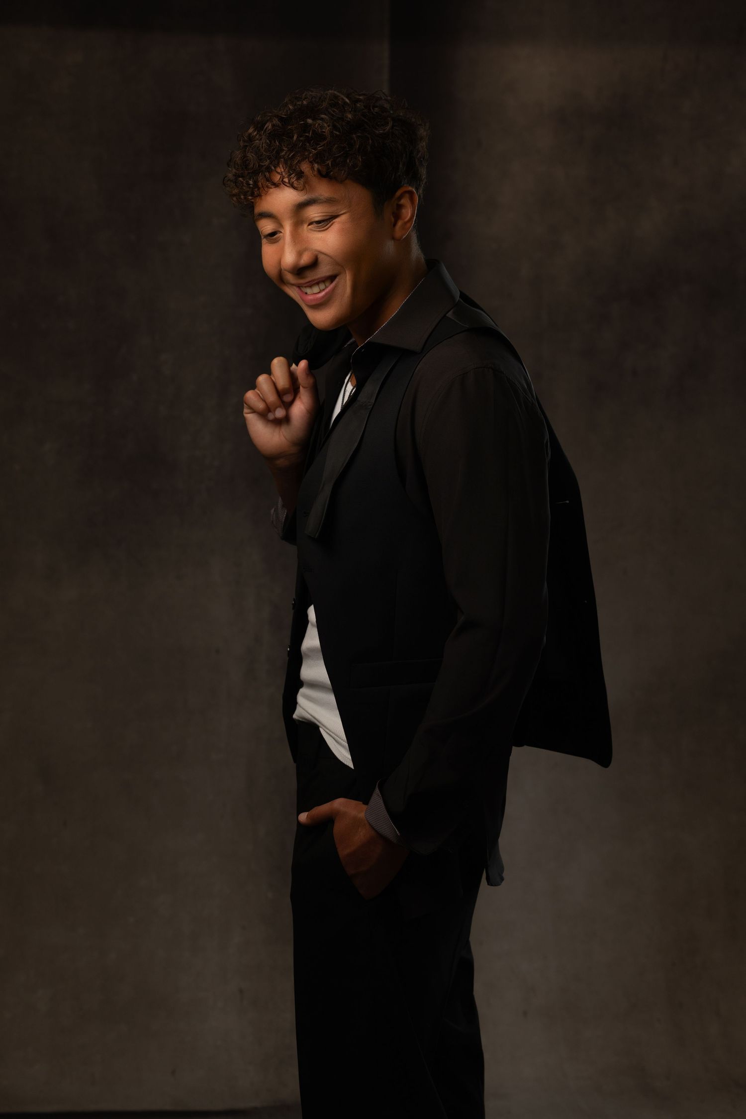 Smiling young man with curly hair wearing a black suit and white shirt, posing in a dark studio setting.