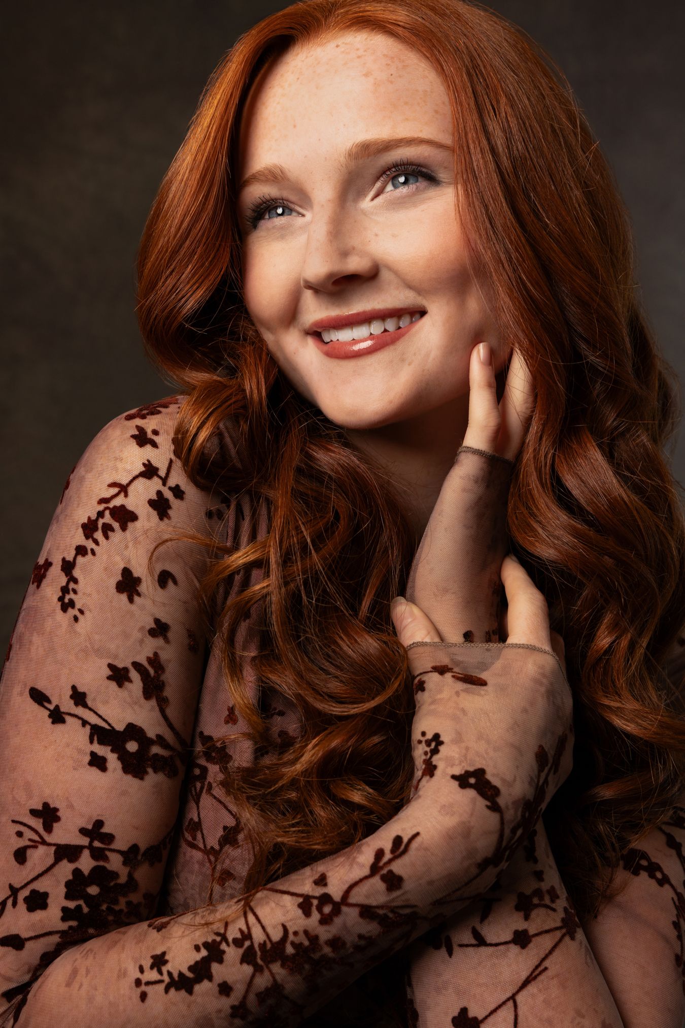 Beautiful redhead woman with freckles in floral lace dress smiling in dark studio portrait.