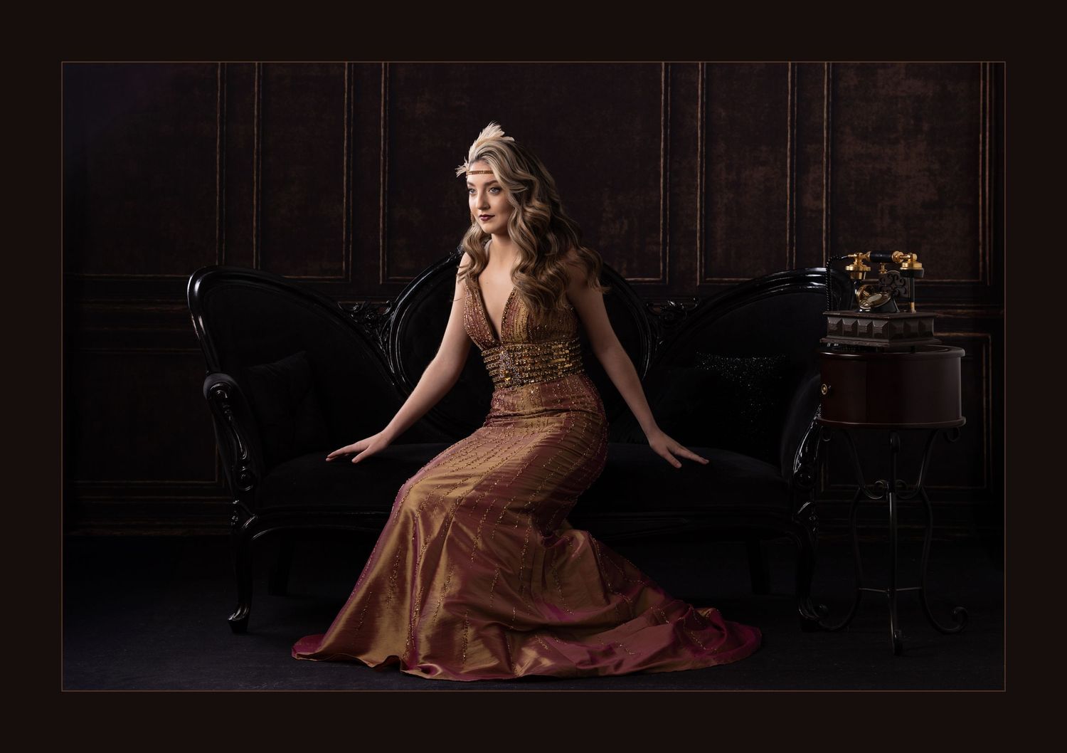 Elegant woman in gold gown sits on black vintage sofa in dark ornate room.