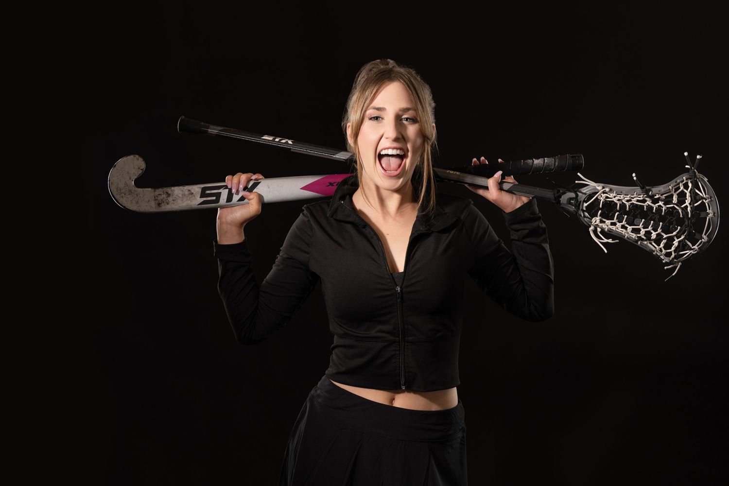 Energetic blonde girl holding field hockey and lacrosse sticks on shoulders against black background.