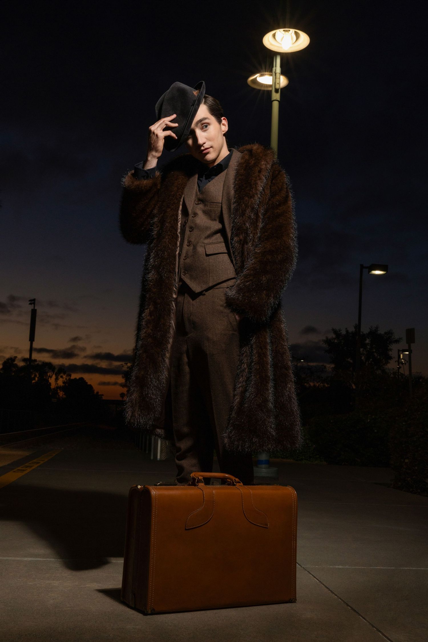 Person in fur coat and fedora hat standing under streetlight at dusk with vintage brown suitcase.