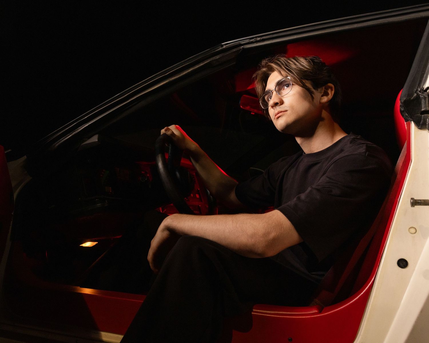 Young man in black shirt sitting in red and white car at night, gripping steering wheel with serious expression.