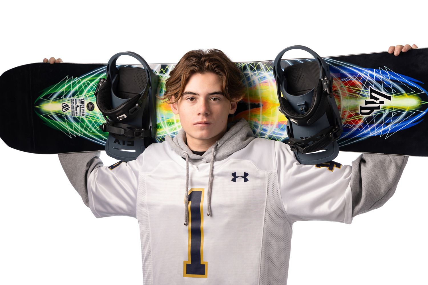 Teen athlete poses with colorful snowboard on shoulders, wearing Under Armour hoodie and number 1 jersey.