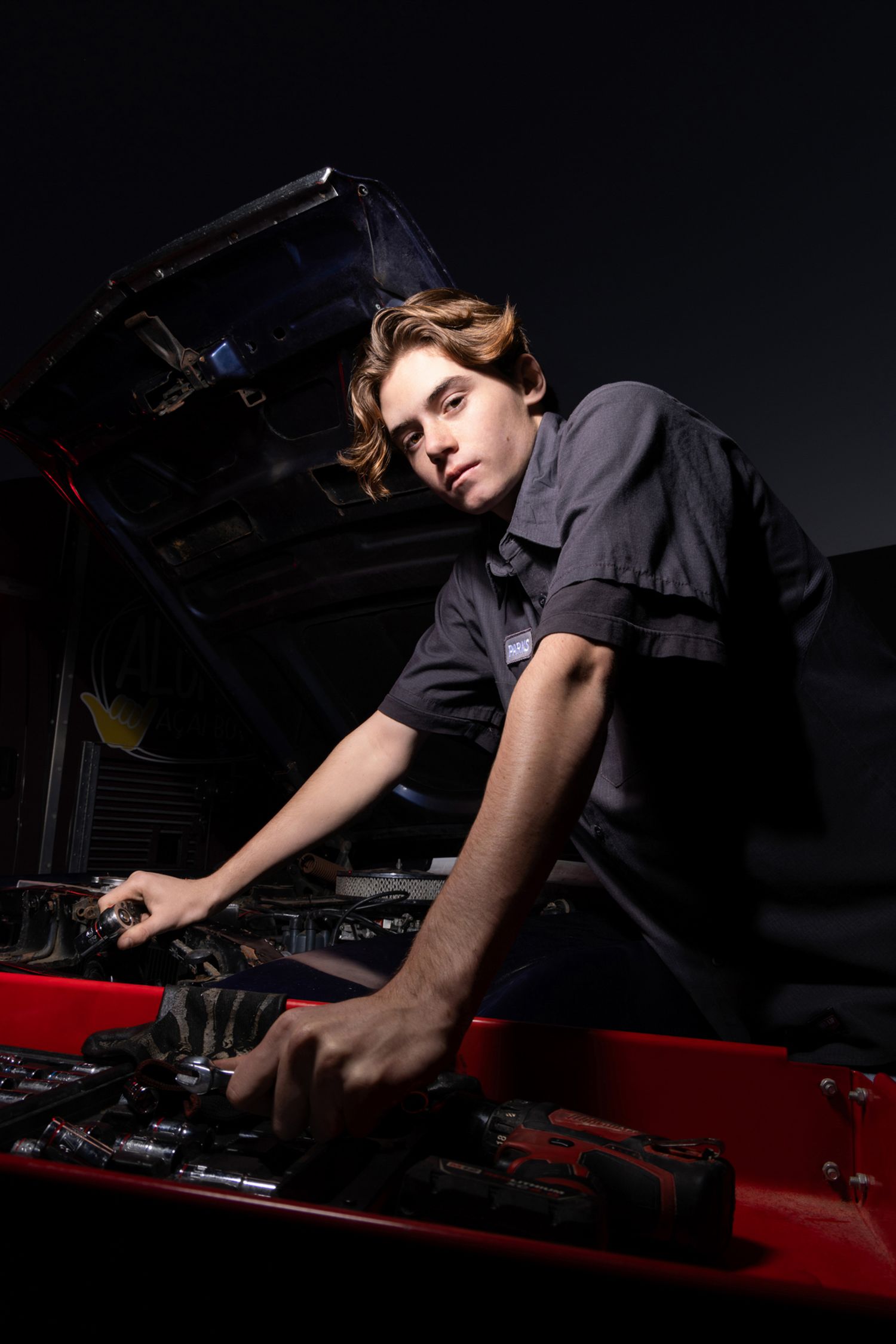 Young mechanic in gray uniform working on car engine with tools at night in dark auto repair shop.