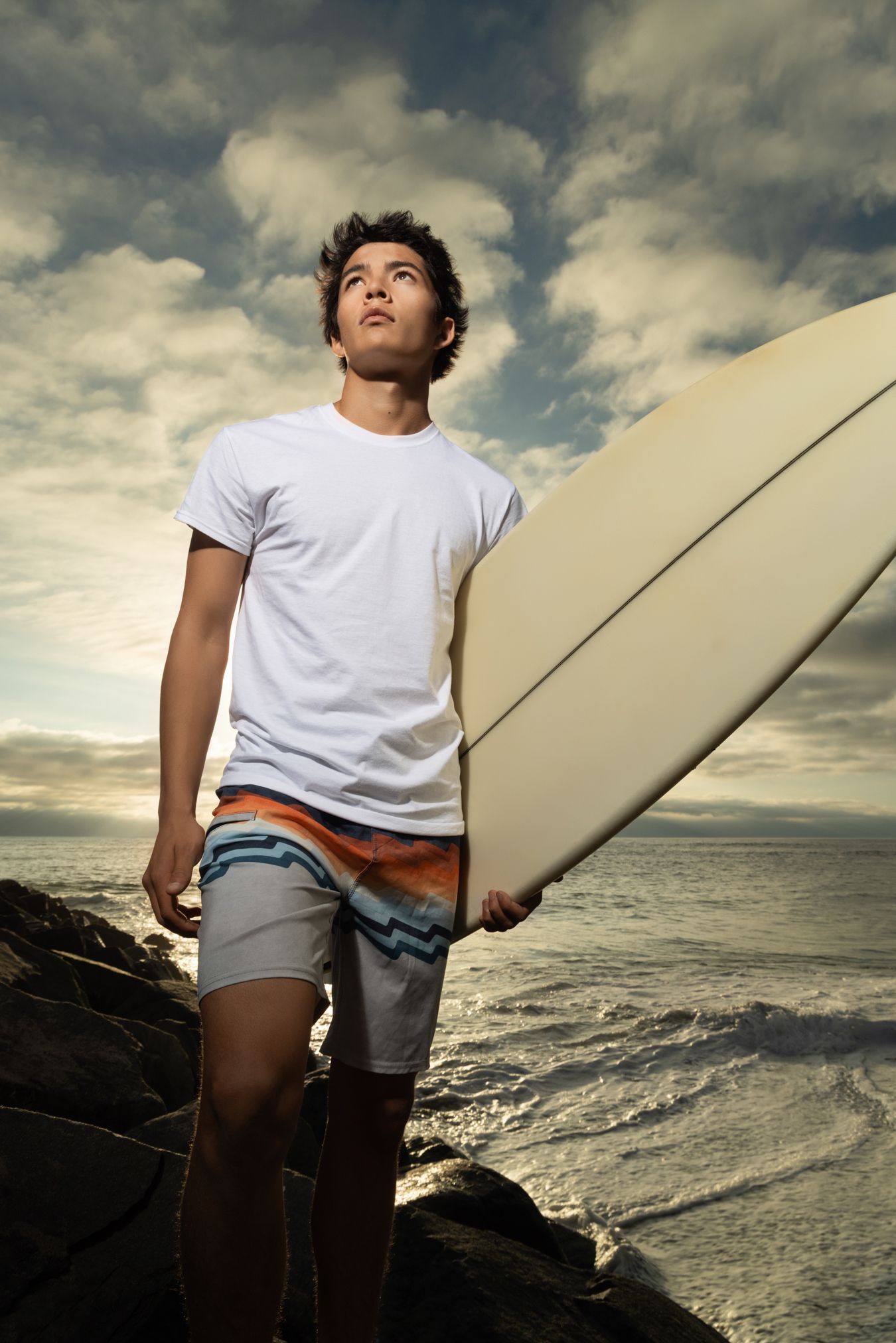 Young male surfer holding a white surfboard on rocky shore, wearing board shorts and white tee, with dramatic cloudy sky.