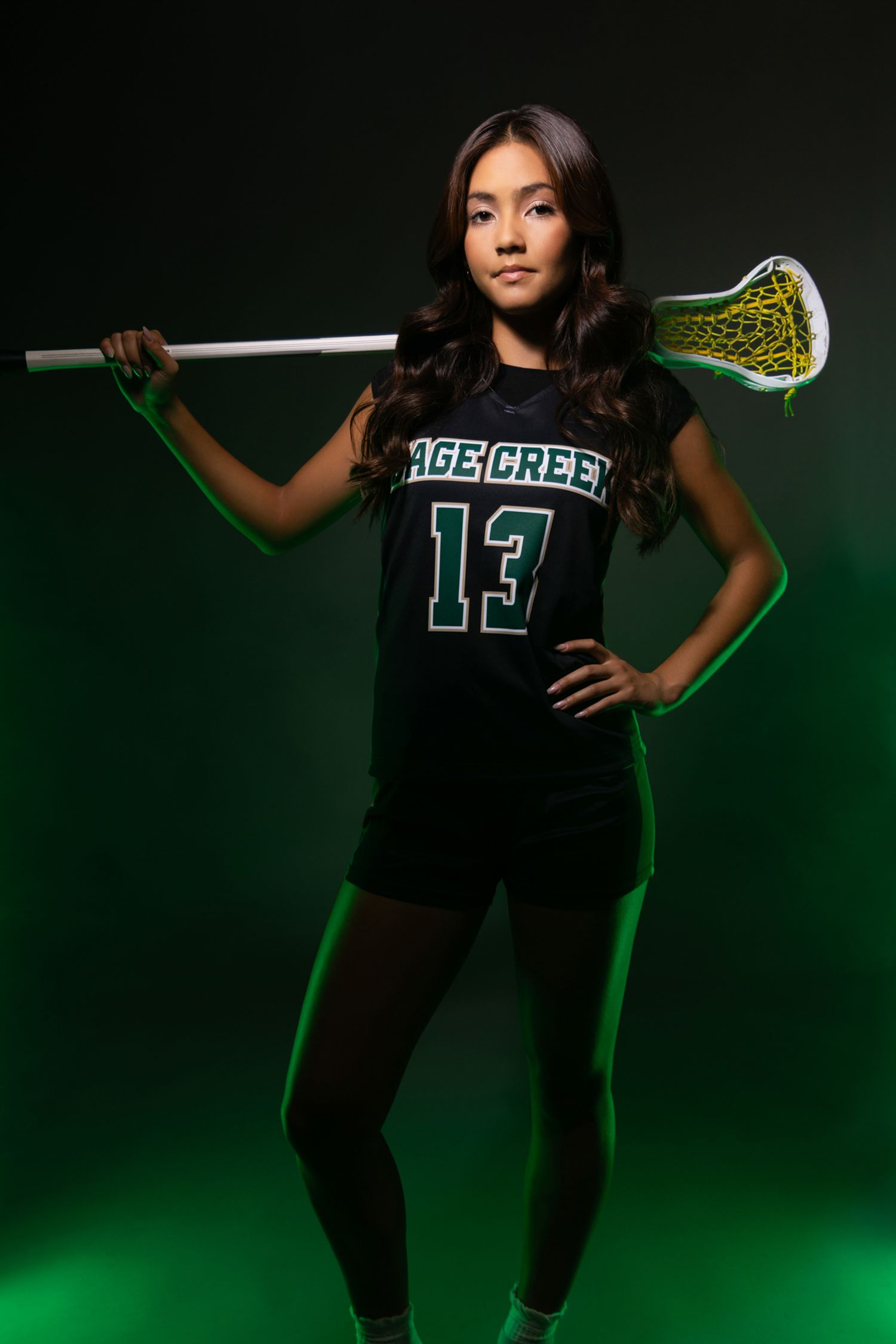 A young female lacrosse player in a green jersey holds a stick over her shoulder in a studio portrait.