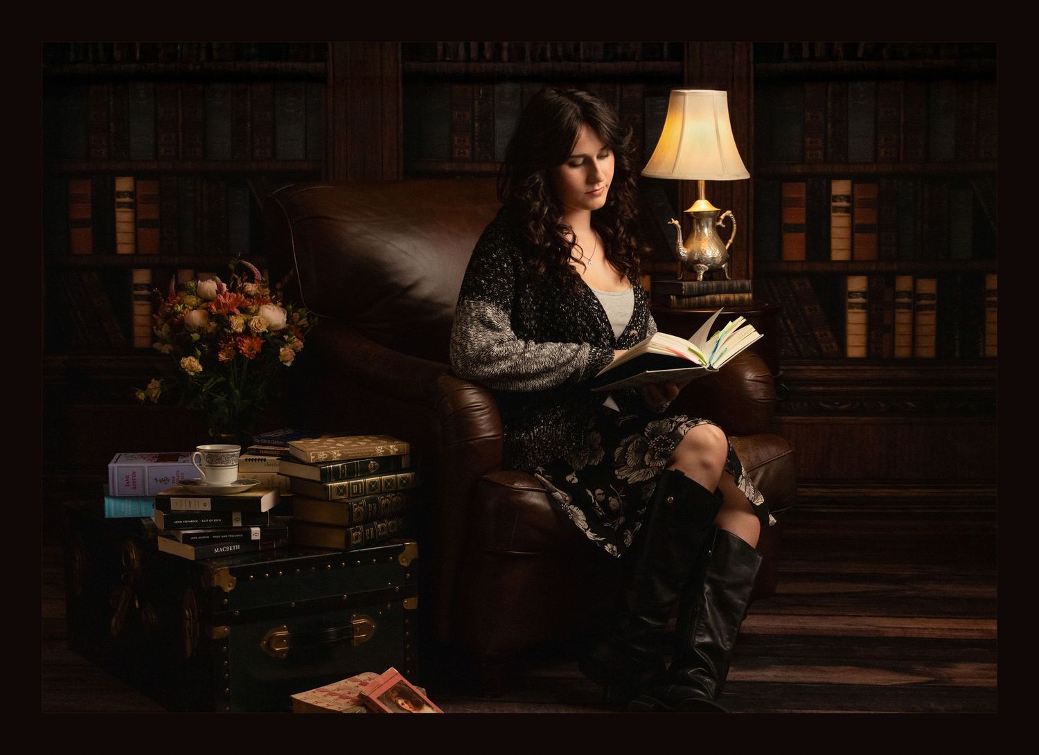 Woman reading a book in a leather armchair surrounded by bookshelves, lit by a warm lamp in a cozy library setting.