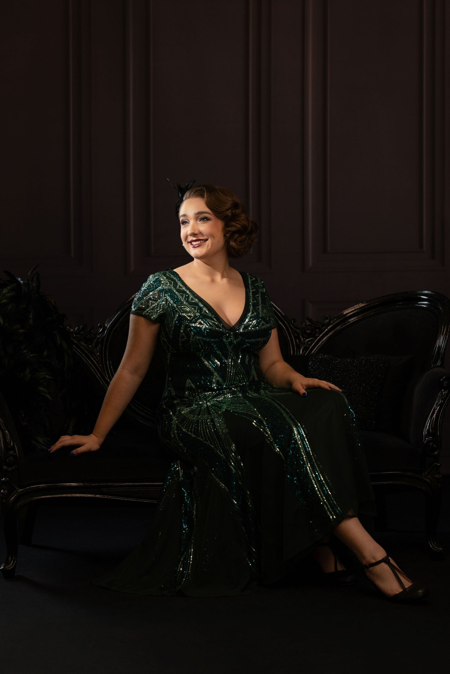 Woman in green sequin jumpsuit seated on dark ornate chair against black paneled wall, elegant studio portrait.