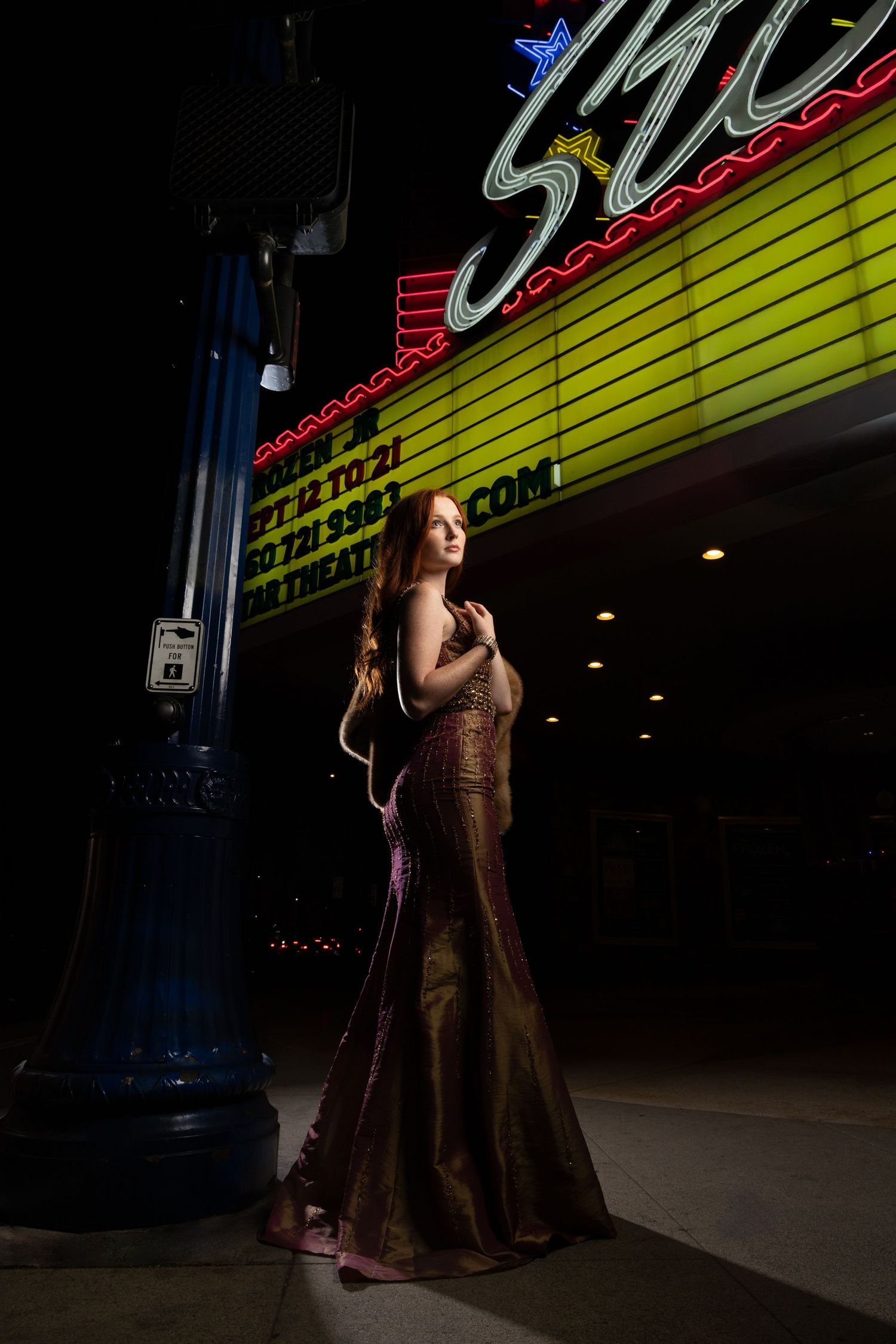 Elegant woman in a gold gown poses under a glowing theater marquee at night.