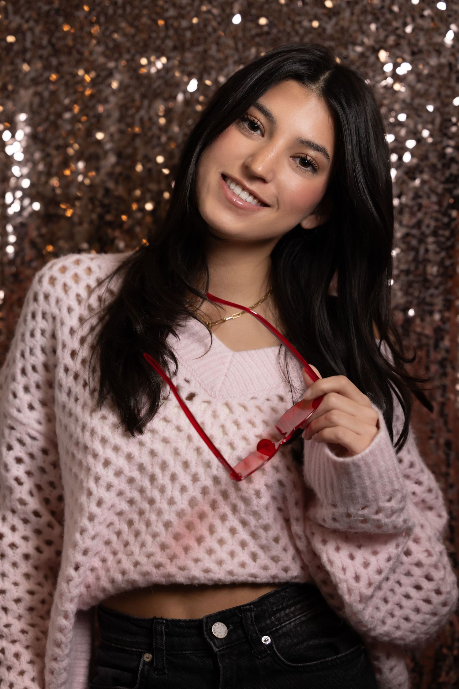 Smiling young woman in pink knit sweater holding red sunglasses against glittery gold sequin backdrop.