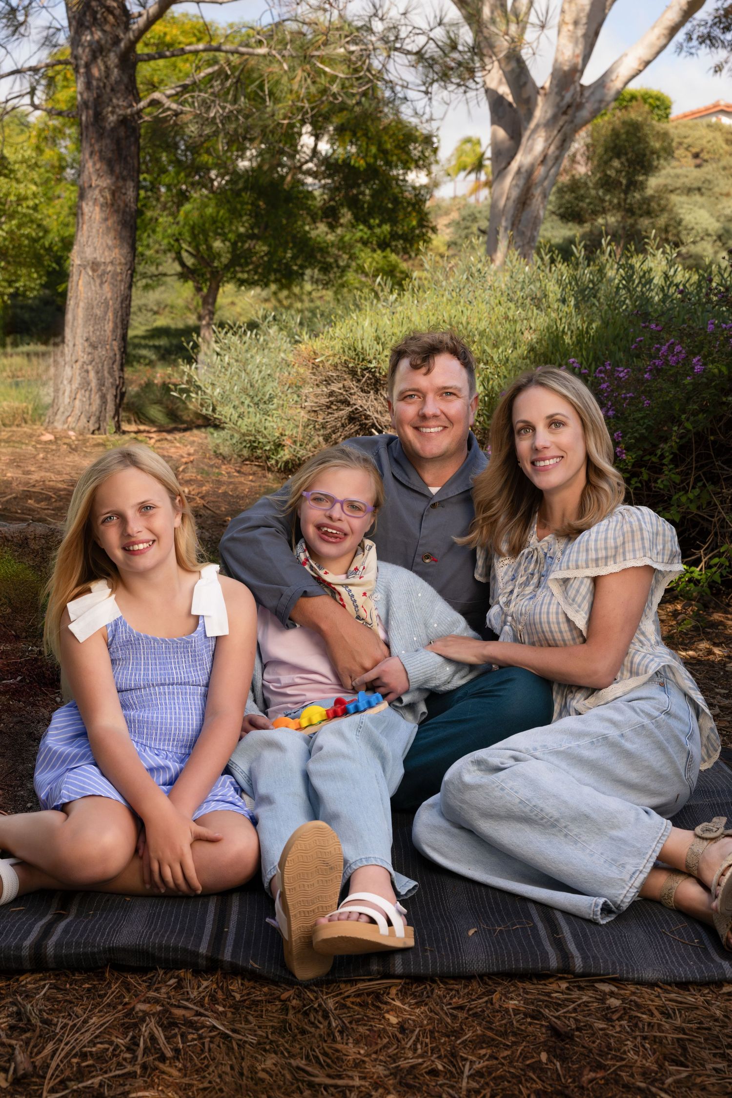 Happy family of four sitting on a blanket outdoors in a lush green park, smiling together.