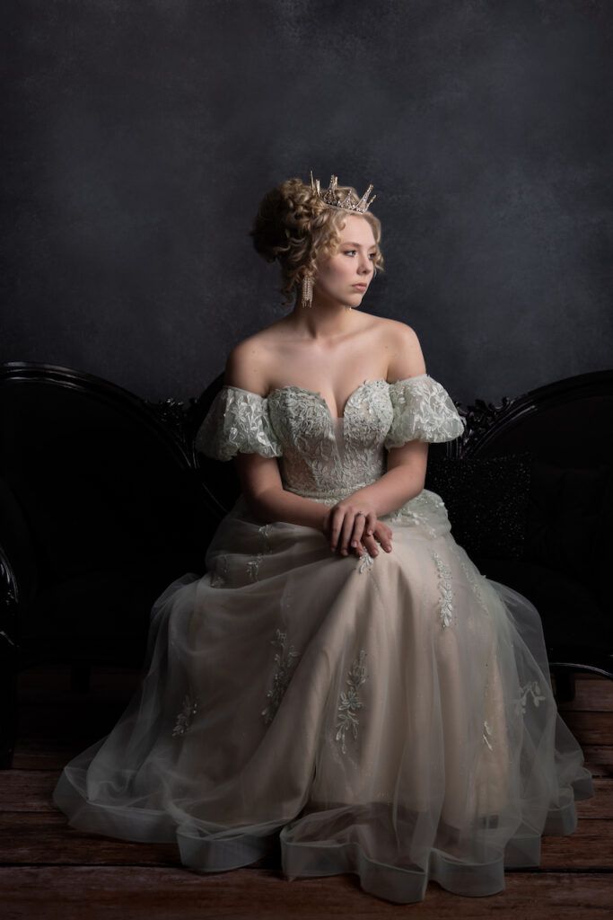 Elegant woman in white off-shoulder ballgown with crown sitting on ornate chair in studio.