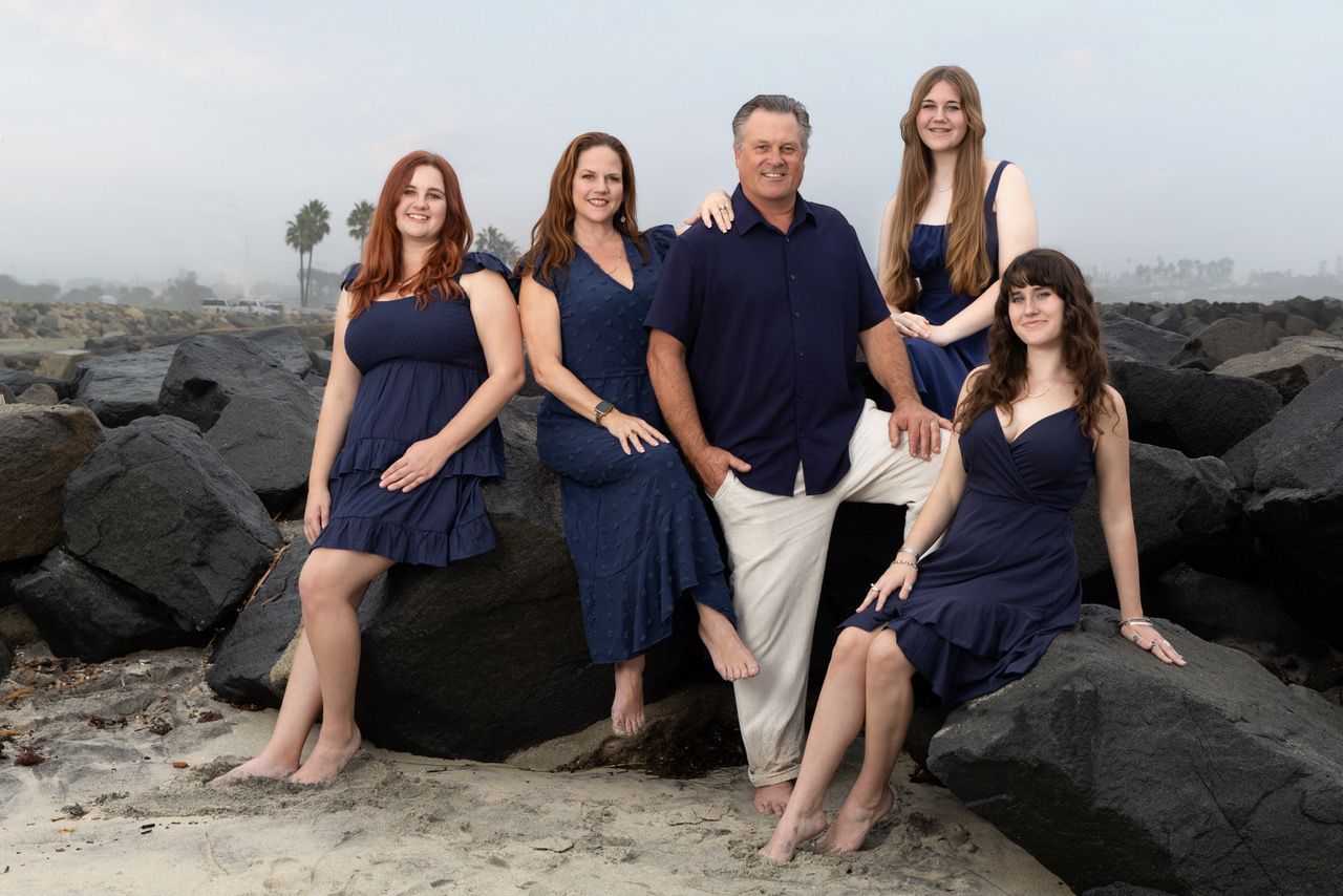 Family of five in coordinating navy outfits posing on rocky beach with ocean and palm trees behind.