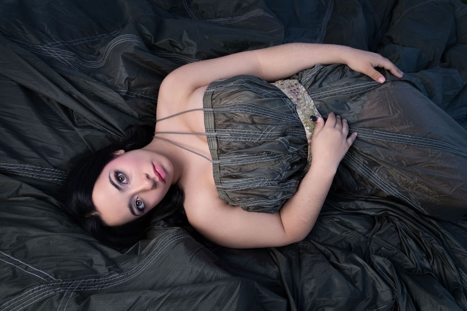 A young woman in a gray strapless gown lies gracefully on dark silk fabric, gazing upward with dramatic makeup.