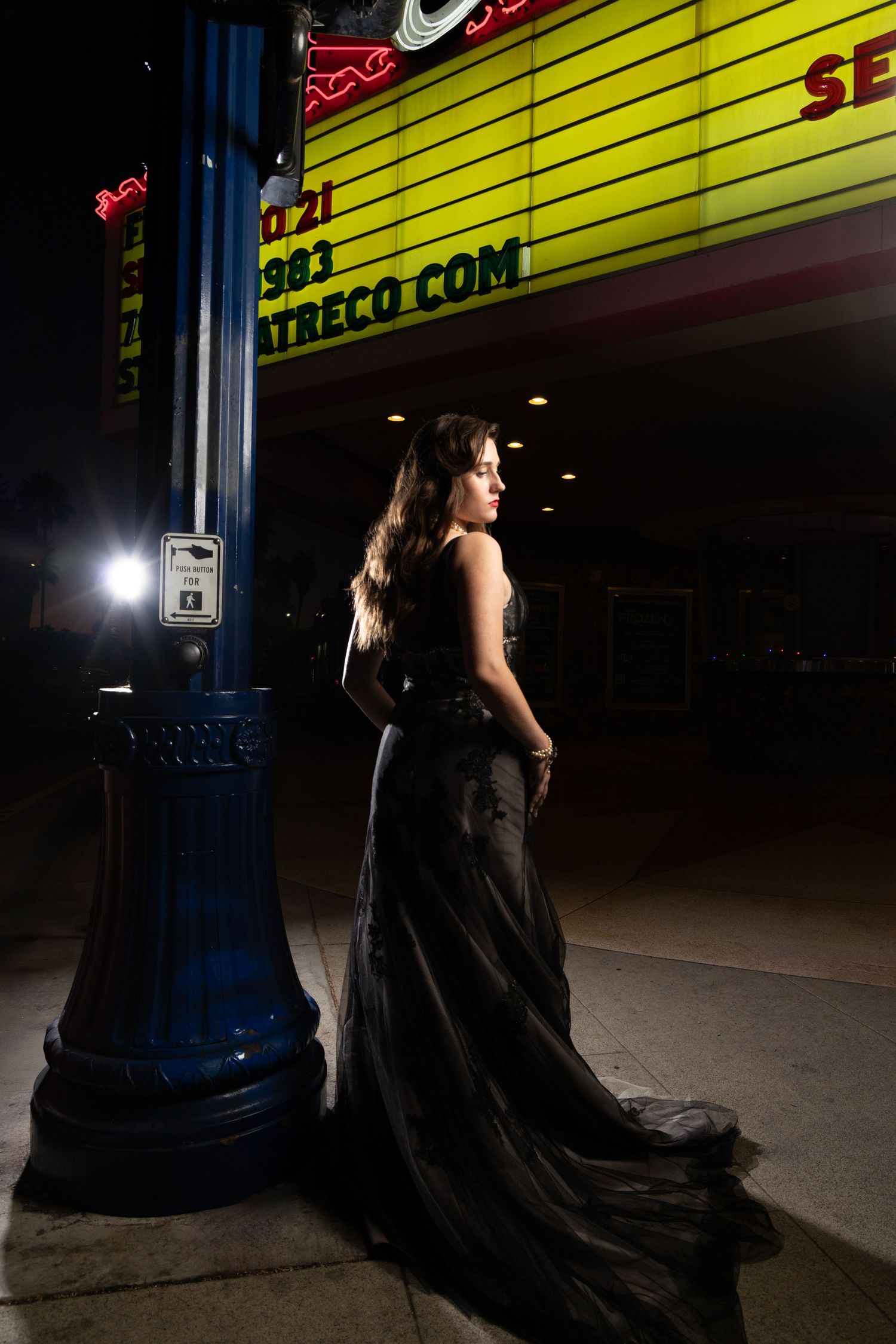 Woman in elegant black gown poses by blue lamppost outside lit theater marquee at night.