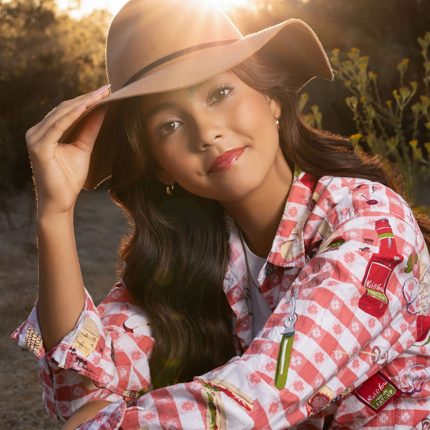 Young woman in cowboy hat and red gingham shirt poses outdoors in golden sunlight.