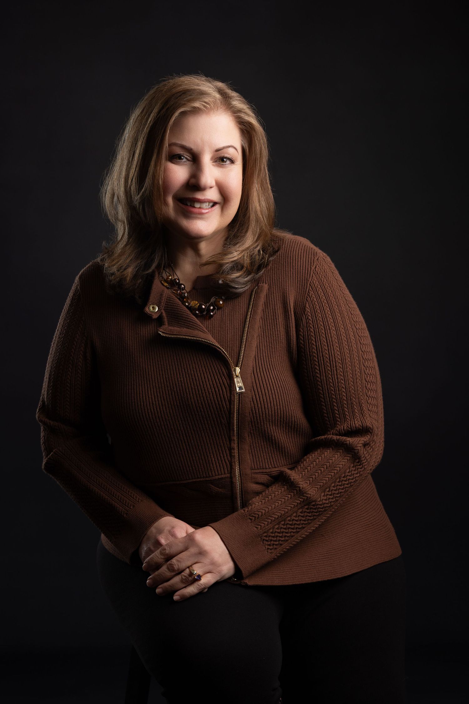 A smiling middle-aged woman with blonde hair wearing a brown zip-up sweater and gold necklace poses against a dark background.