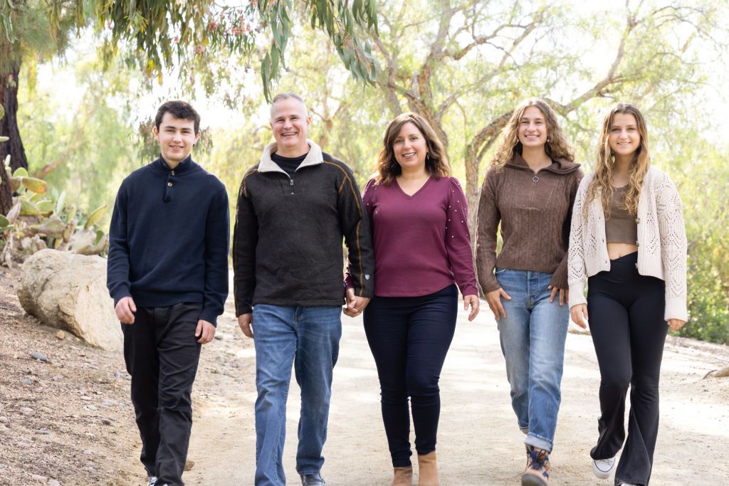 Family of five smiling and walking together on a scenic outdoor path surrounded by lush green trees.