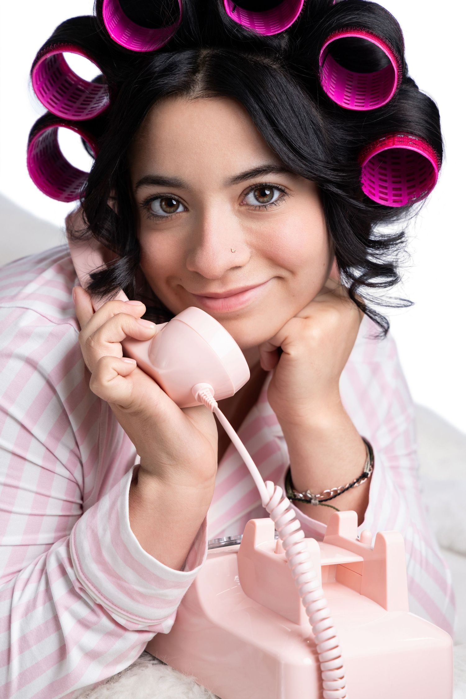 Young woman with pink hair rollers smiling while talking on a vintage pink rotary telephone in striped pajamas.