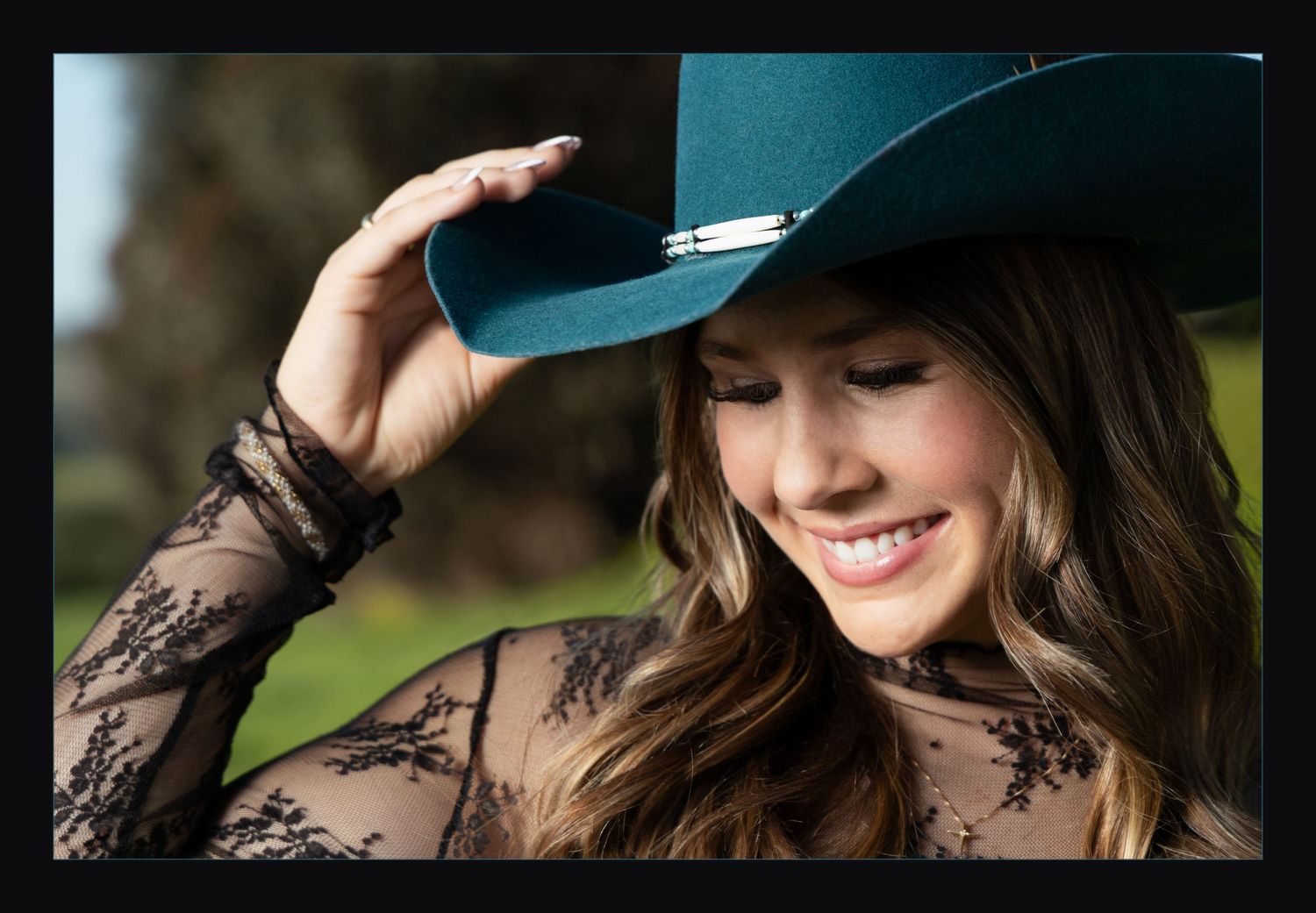 Smiling woman in teal cowboy hat and lace top looks down in soft outdoor light.