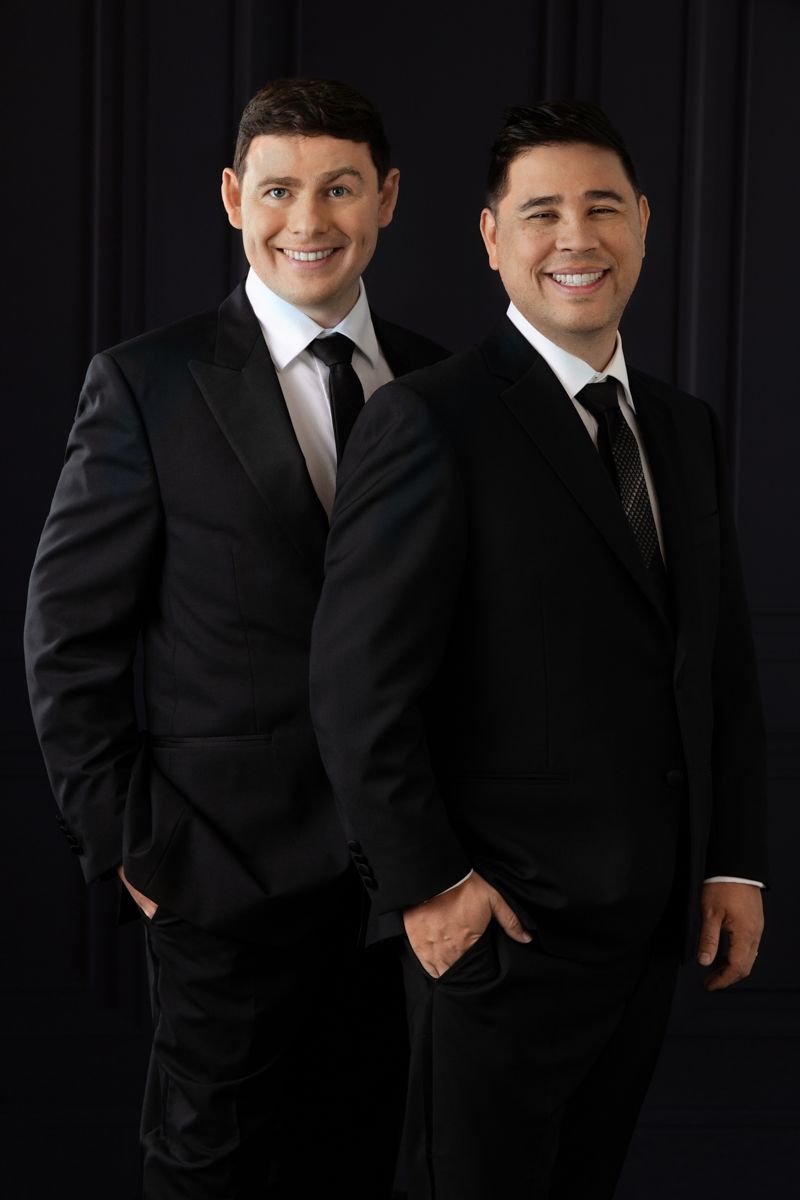 Two men in matching black suits and ties smiling confidently in a professional portrait against dark background.