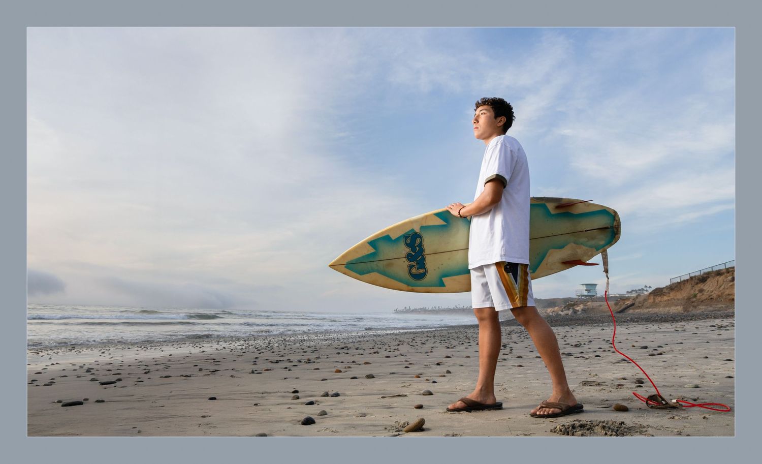 Young boy holding a Glide surfboard on a sandy beach, gazing at ocean waves during golden hour.