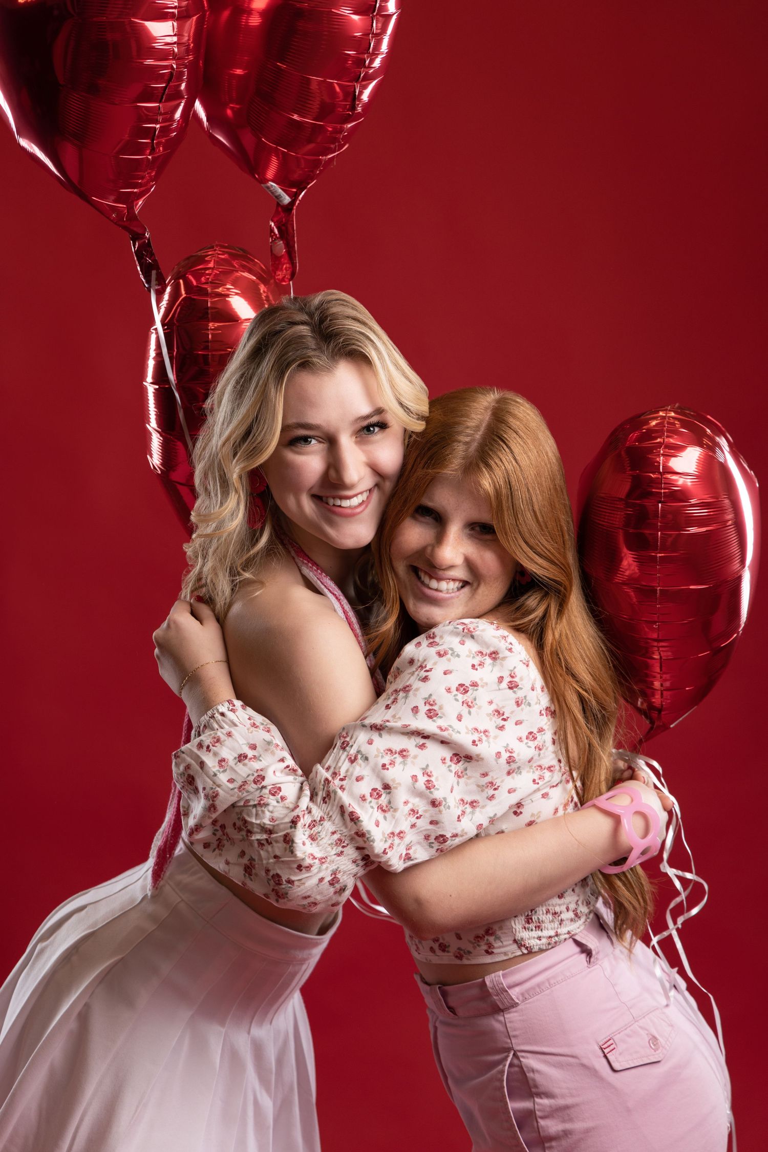 Two smiling women embrace holding red heart balloons against vibrant red background.