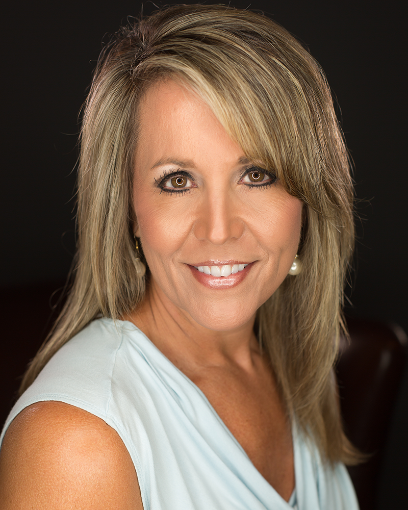 Professional headshot of smiling blonde woman in light blue top against dark background.