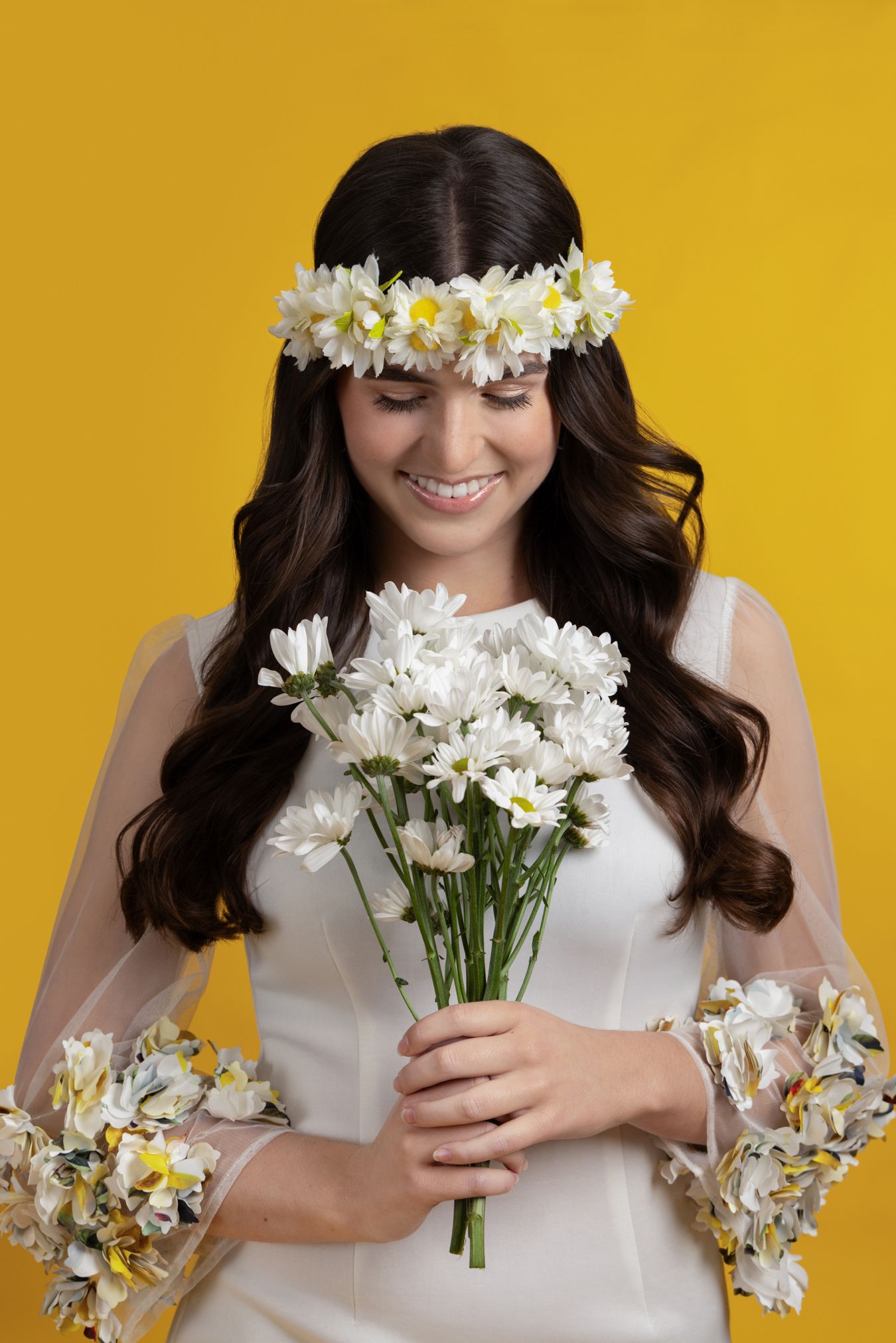 Smiling brunette bride in white dress holding white daisy bouquet with matching floral crown on yellow background.