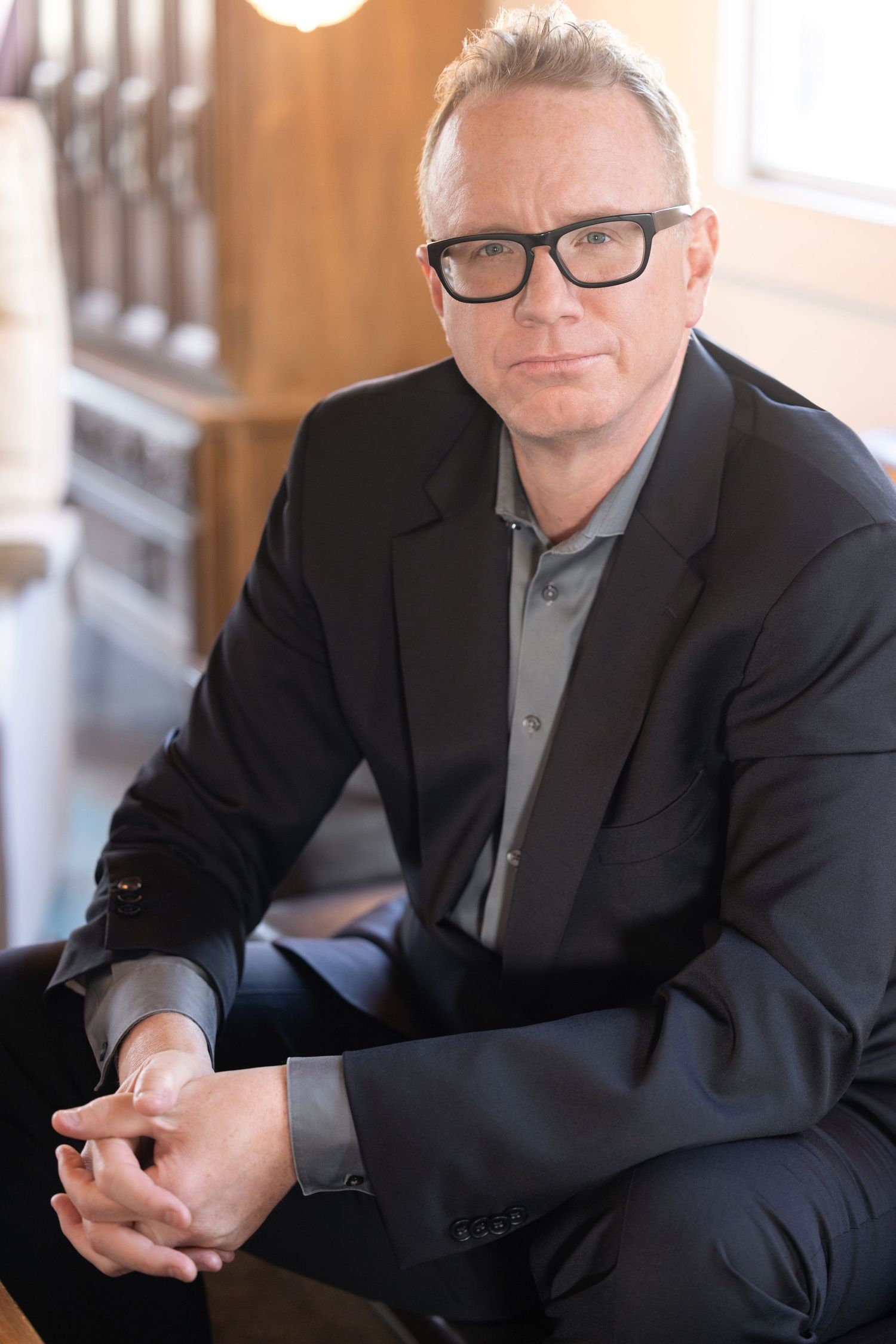 Professional man in black blazer and glasses sitting with clasped hands in a warmly lit interior setting.