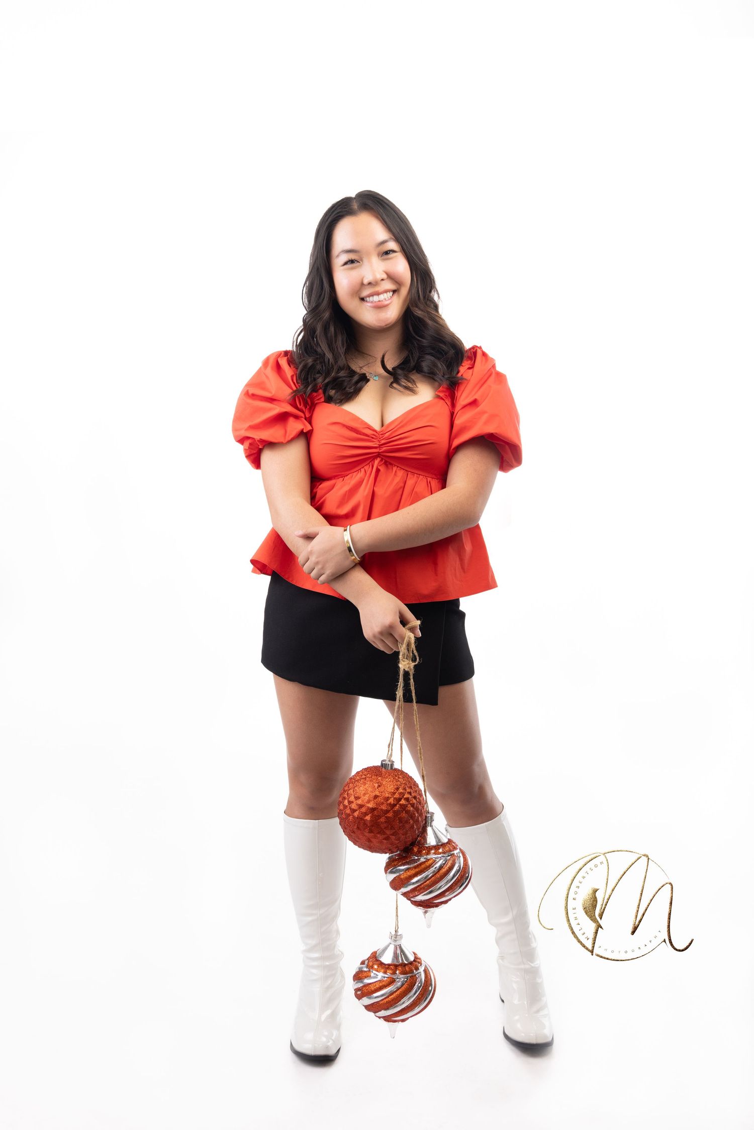 Young woman in red top, black skirt, and white boots holding Christmas ornaments, smiling against white background.