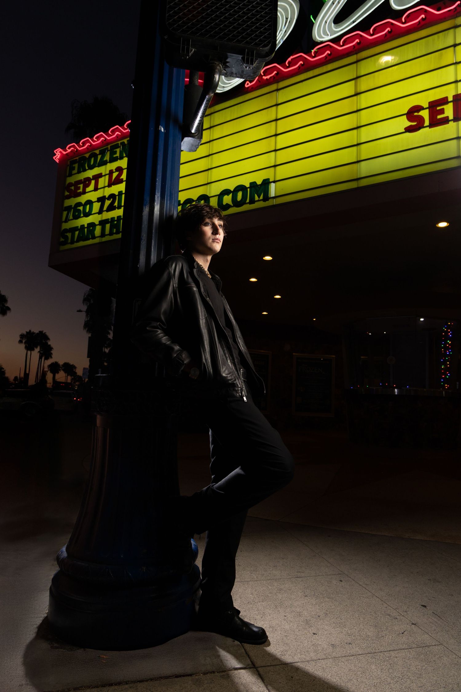 Young man in black leather jacket leaning against a pole outside a illuminated theater marquee at dusk.