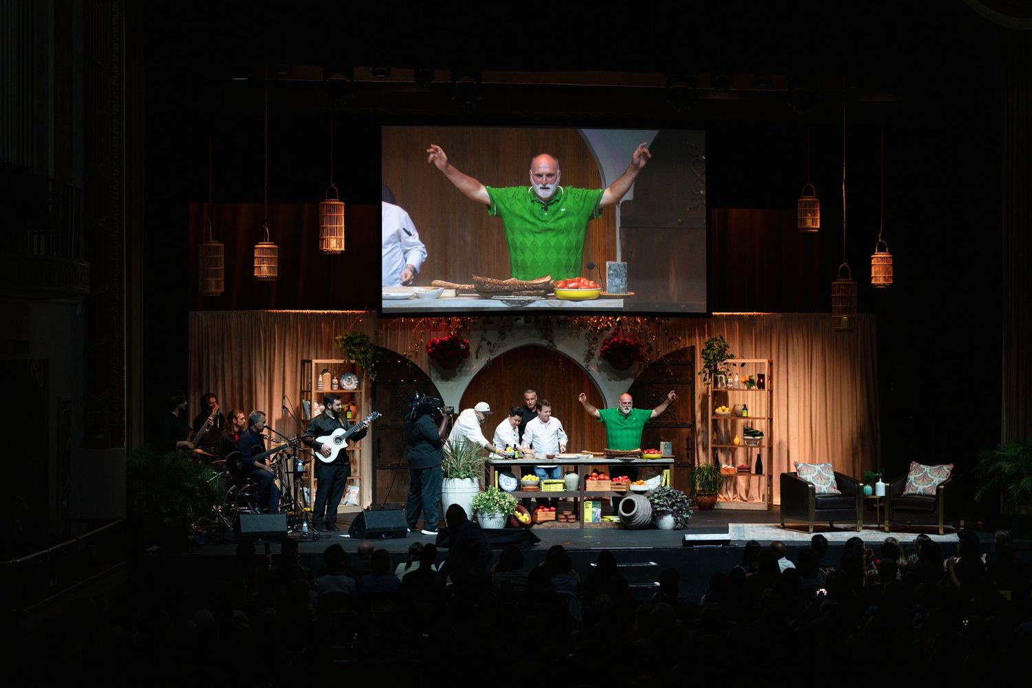 Live cooking show on stage with chef in green shirt on screen, band, and audience in dark venue.