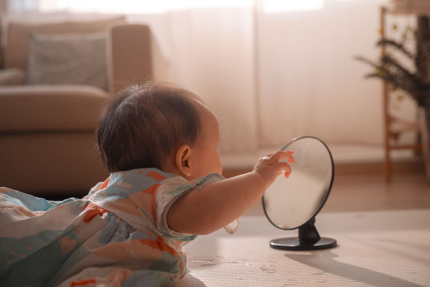 A curious baby reaches toward a small round mirror on the floor in a warm, sunlit living room.