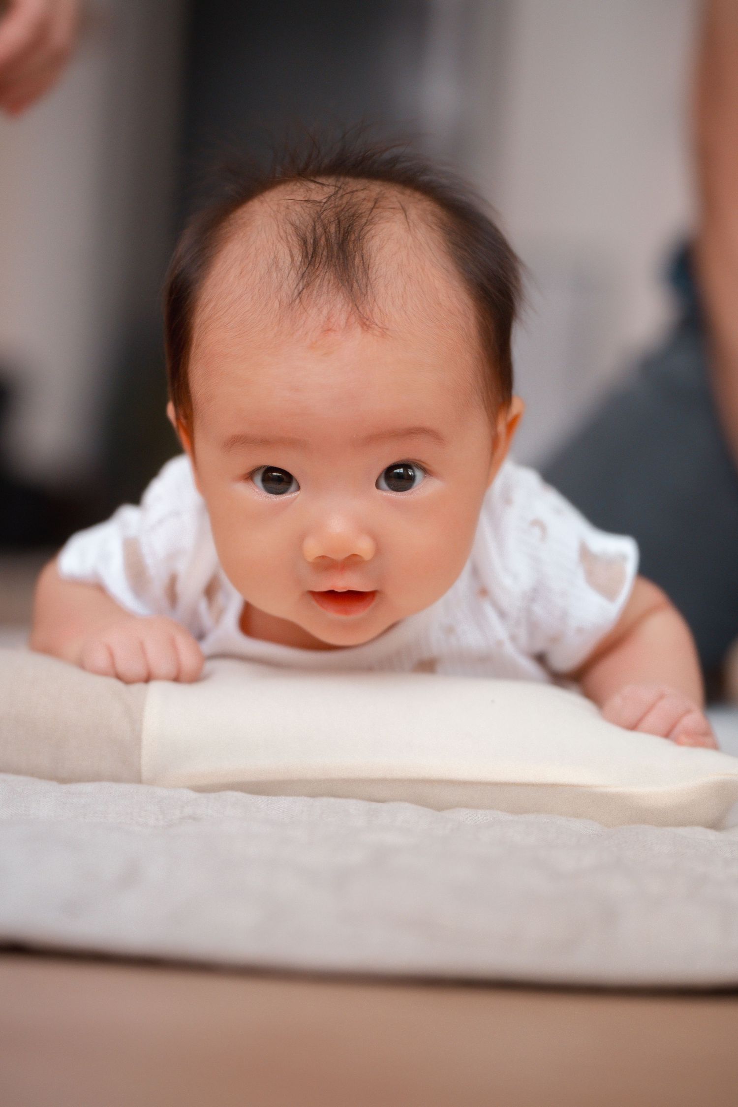 A baby in a white outfit peeks over the edge of a surface with a curious expression.