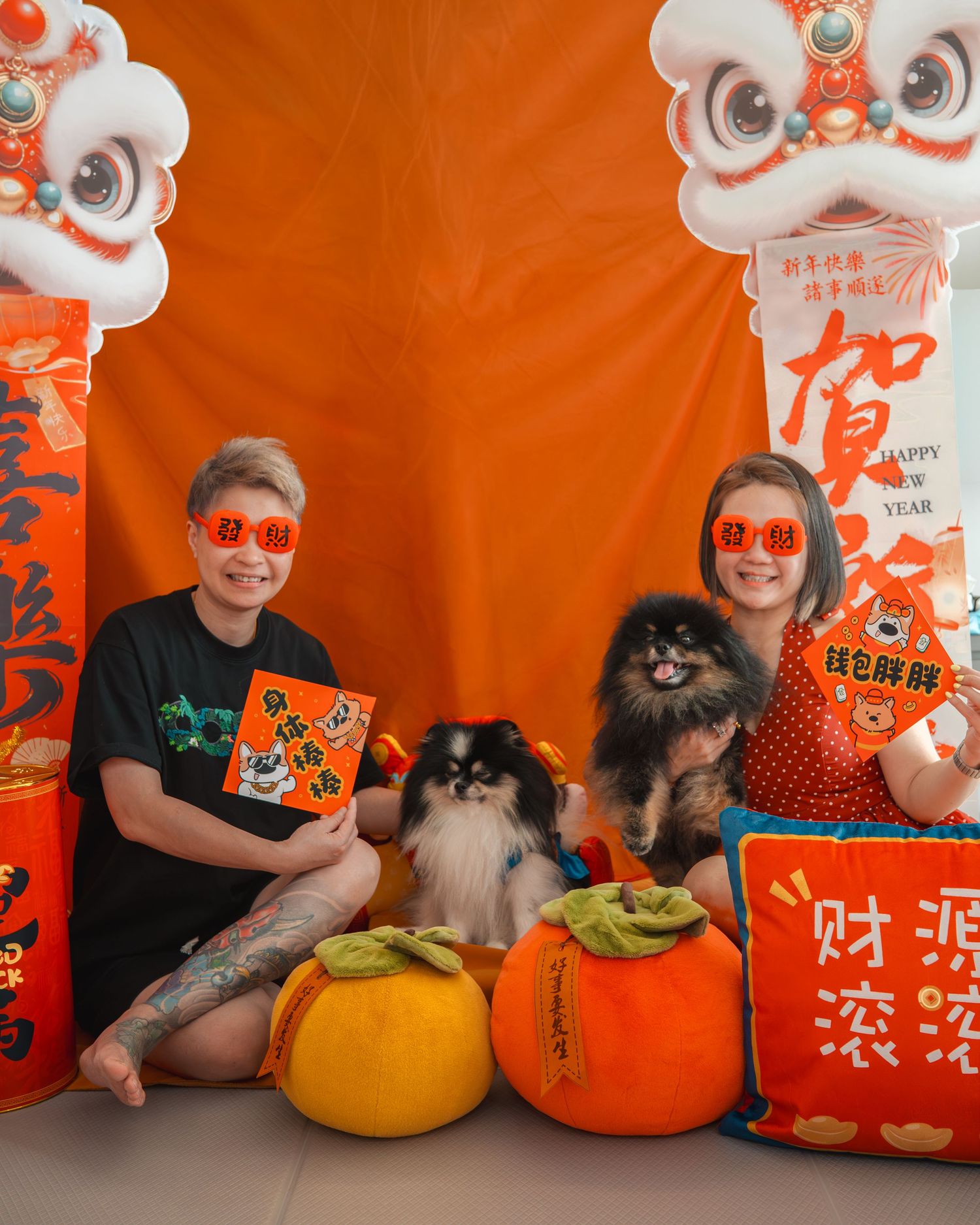Two people and two Pomeranian dogs pose before a vibrant Chinese New Year backdrop with lion dancers and festive decorations.