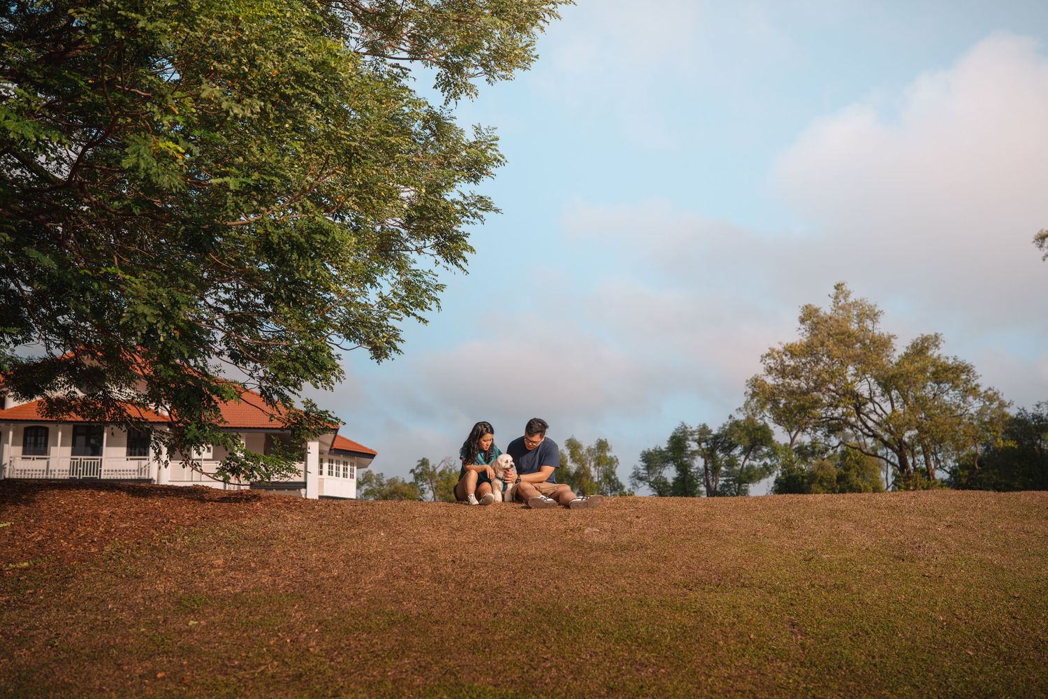 Couple sitting together on a grassy hill near a white colonial building under a blue sky with trees.
