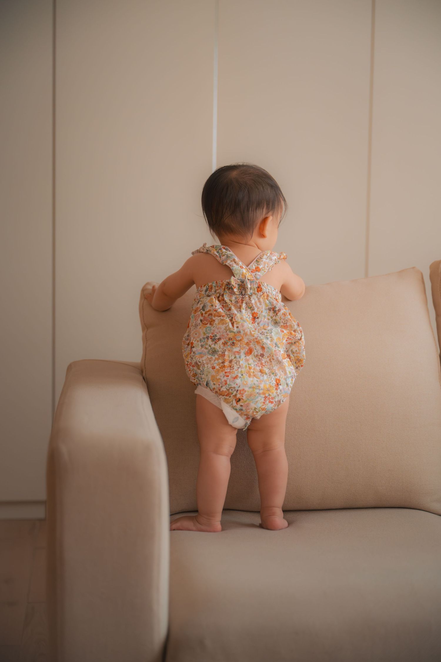 Baby in floral romper standing on beige sofa, viewed from behind.