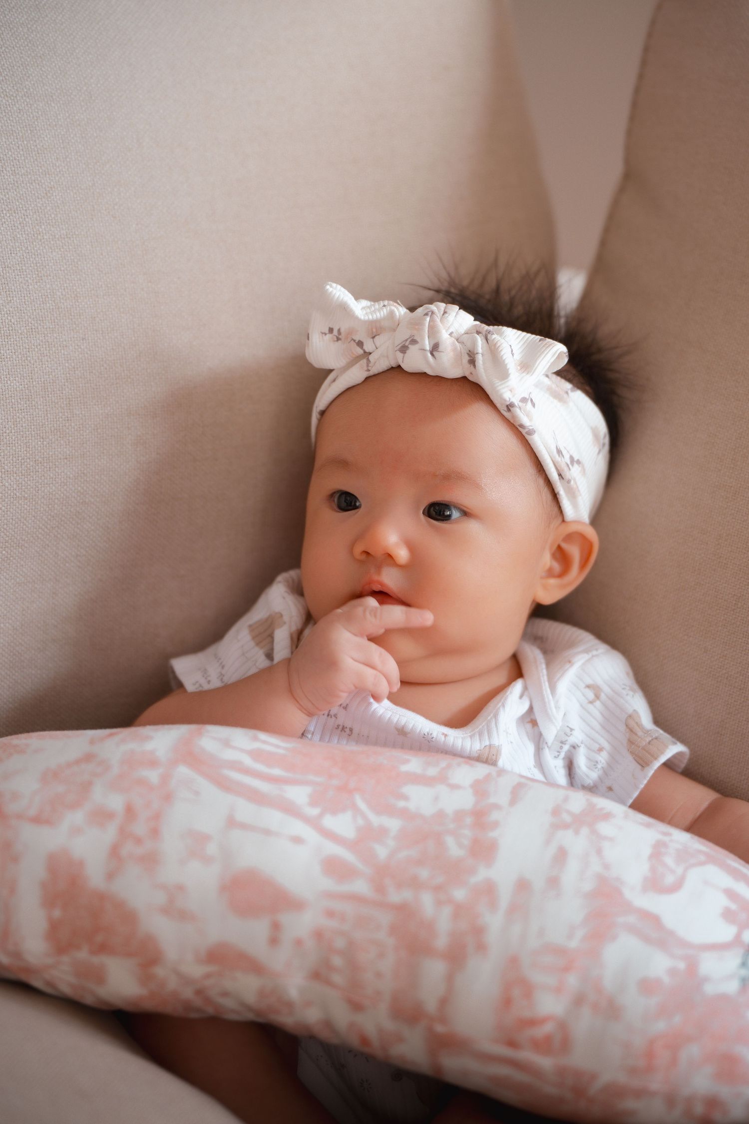 Baby wearing white headband and shirt lies on couch with pink patterned pillow.