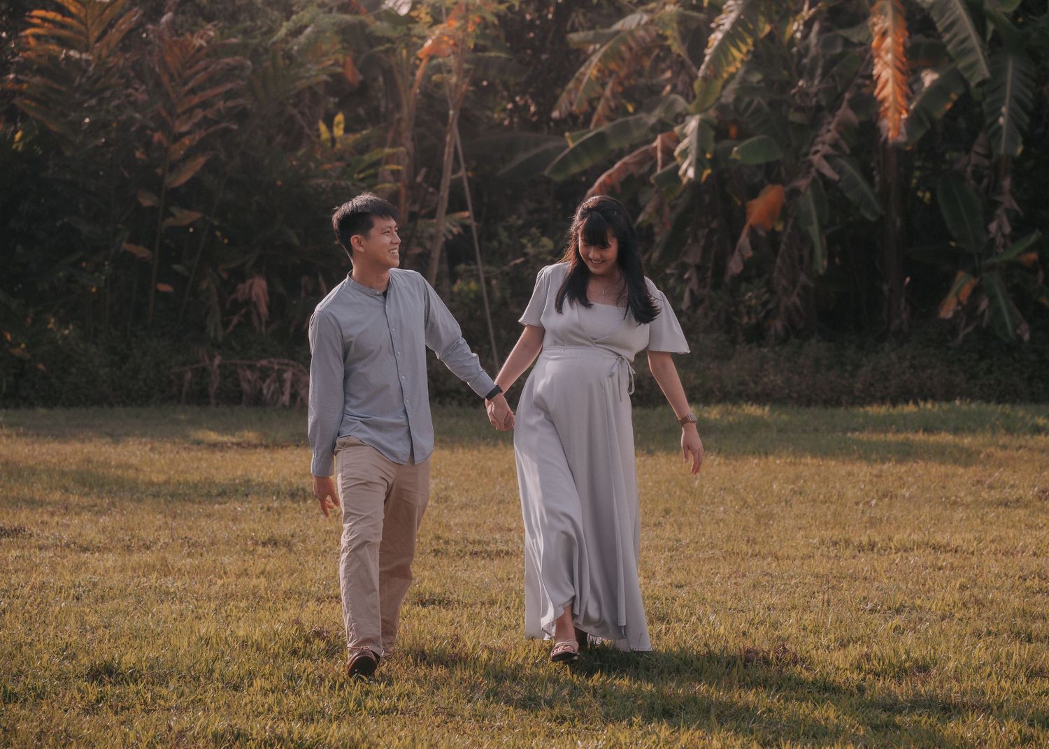 Couple holding hands walking on green grass field with tropical palm trees in golden sunset light.