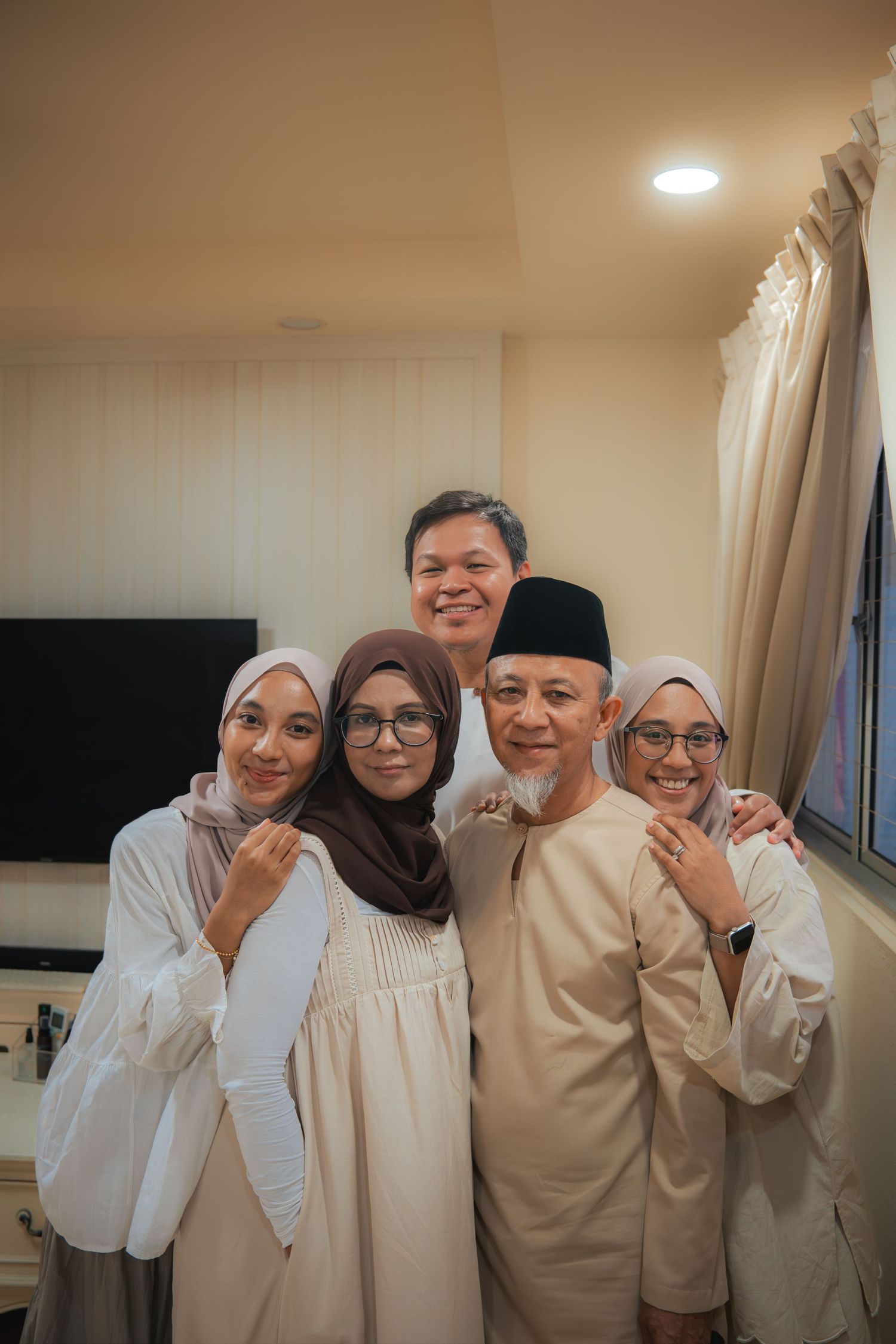 Happy Muslim family of five posing together indoors, wearing traditional white attire and hijabs, smiling warmly.