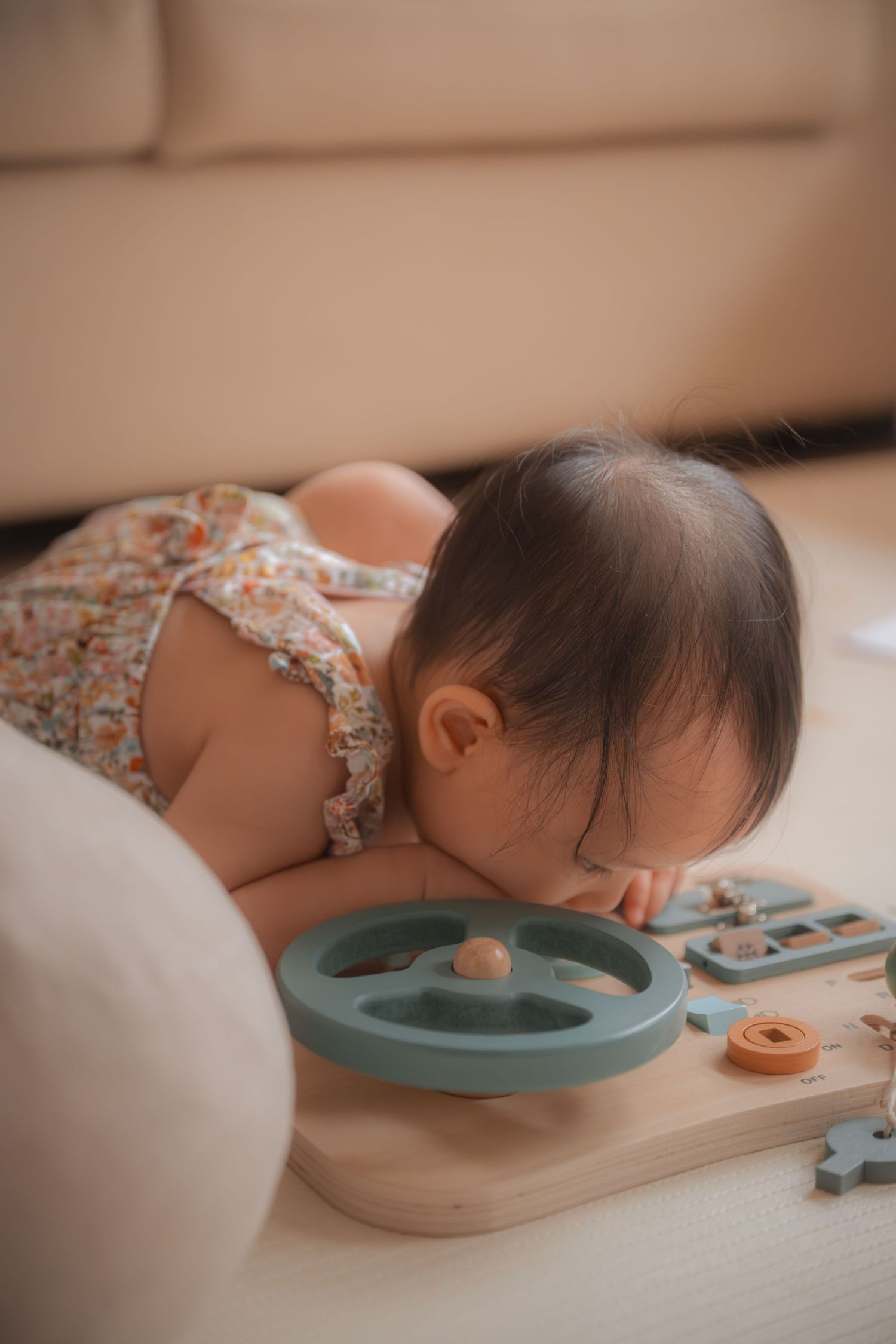 Baby in floral romper leaning over colorful wooden shape-sorting toy on floor near a couch.