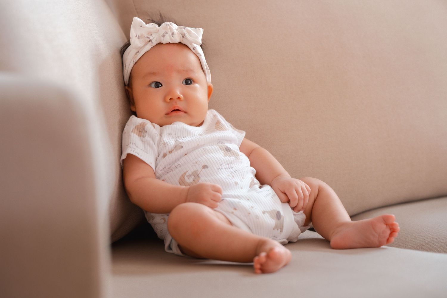 A baby wearing a white outfit and headband lies on a beige couch in a series of connected photos.