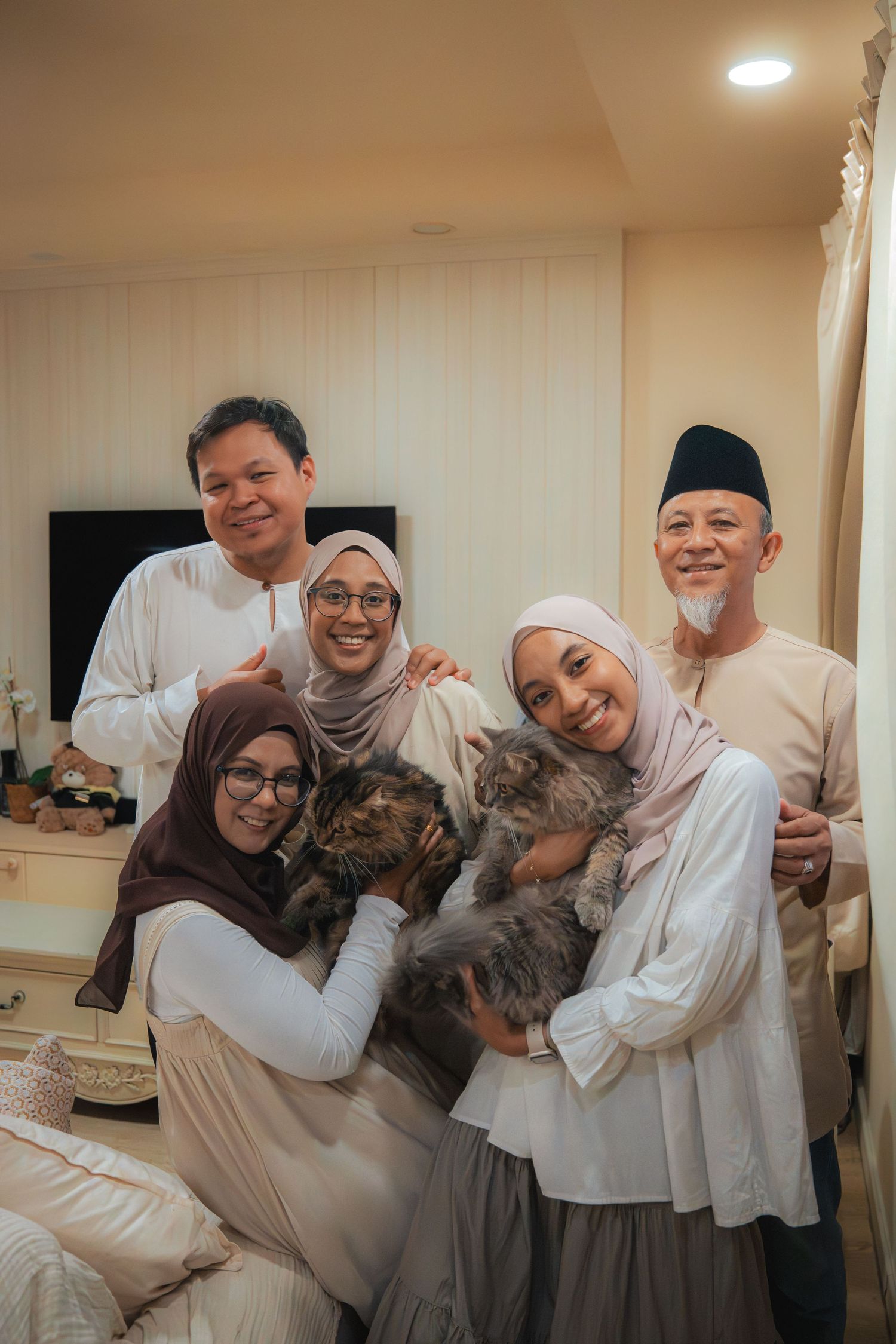 Muslim family in traditional attire smiling together while holding fluffy cats in a cozy living room.