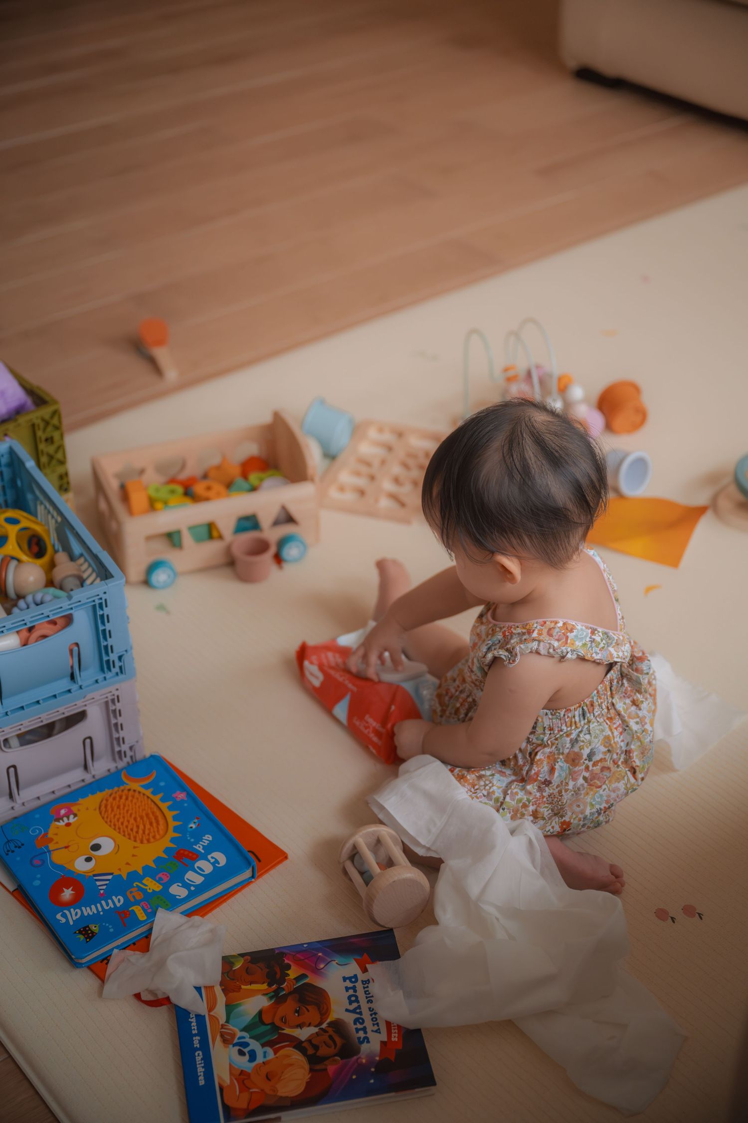 Baby girl in floral romper playing with wooden toys and colorful books on a soft play mat.